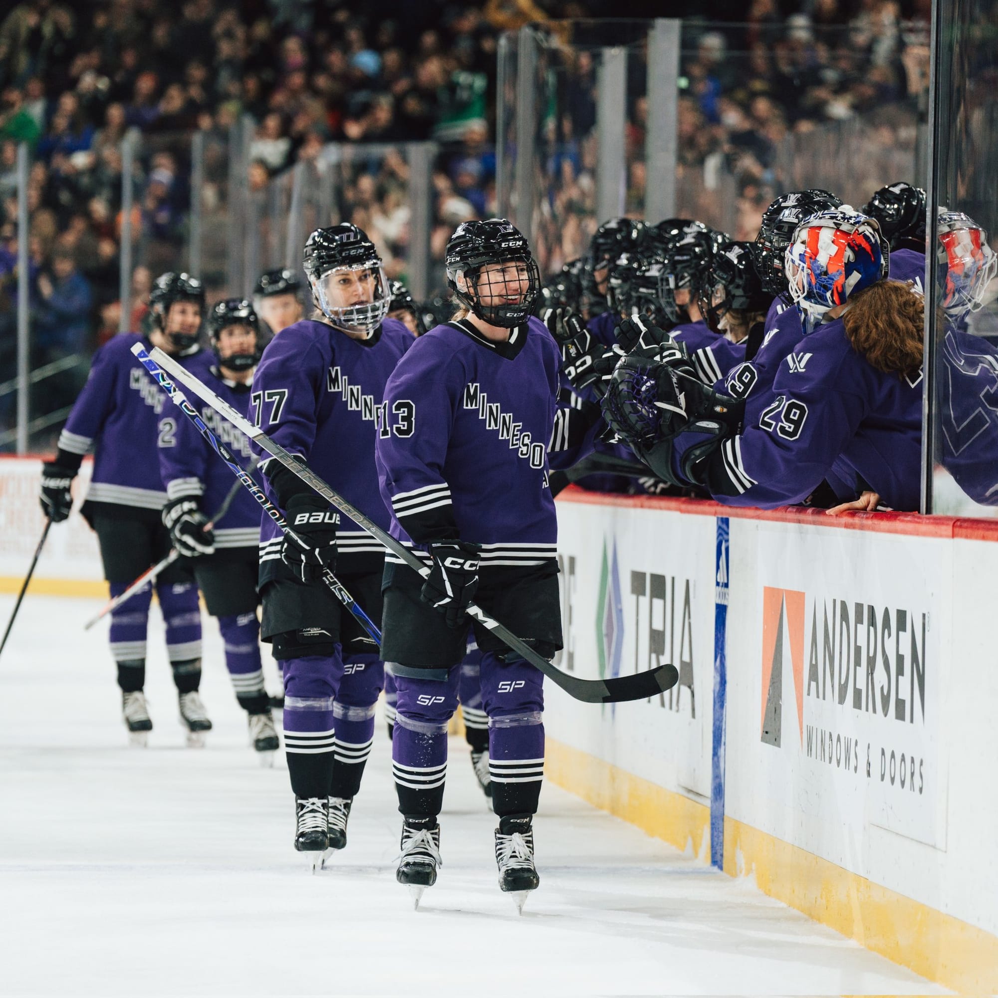 Grace Zumwinkle and her teammates celebrate a goal with a fist-bump line at the bench in their purple home uniforms..