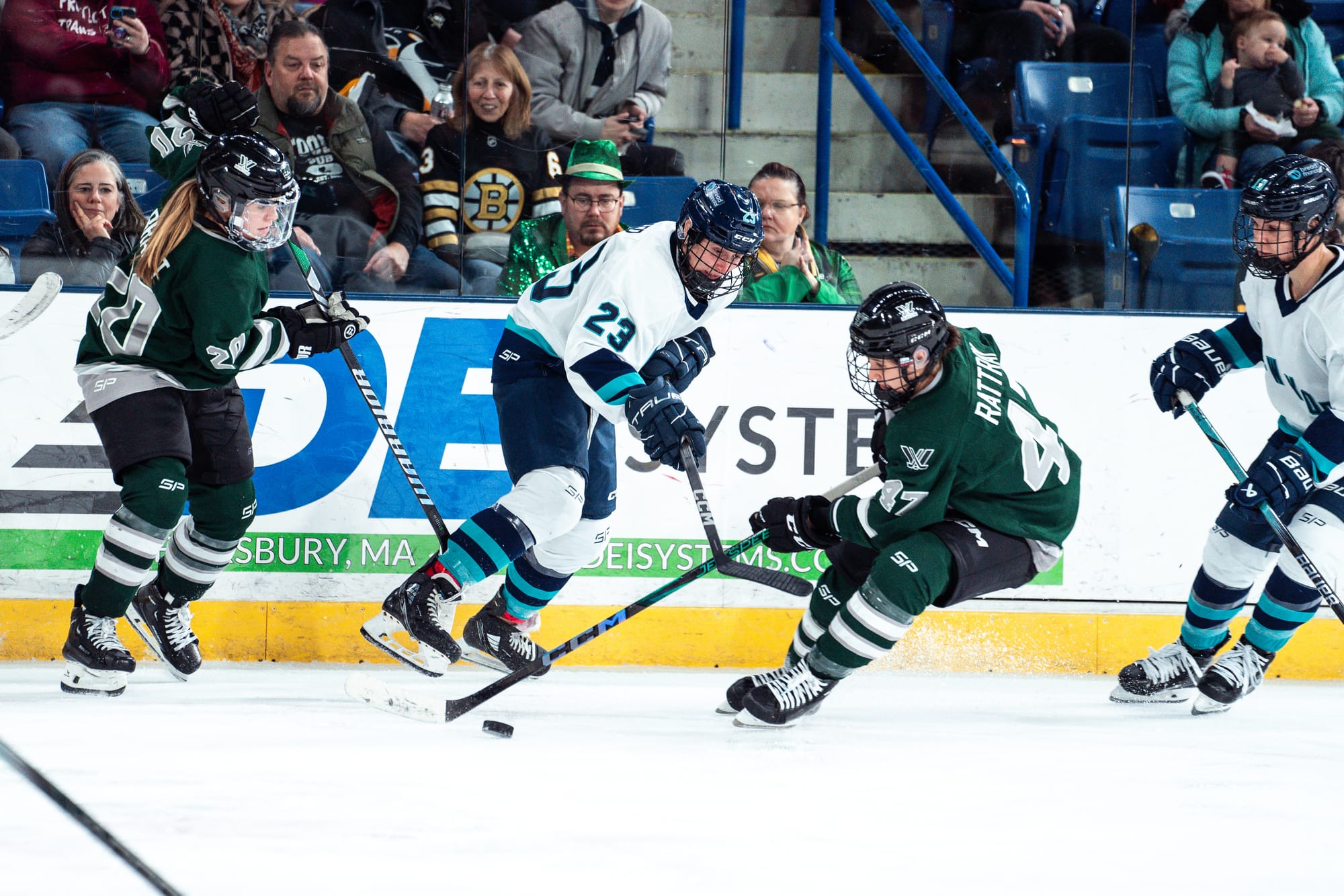 Jamie Lee Rattray, wearing a green home uniform, skates with the puck under pressure from New York skaters, wearing white away uniforms. 