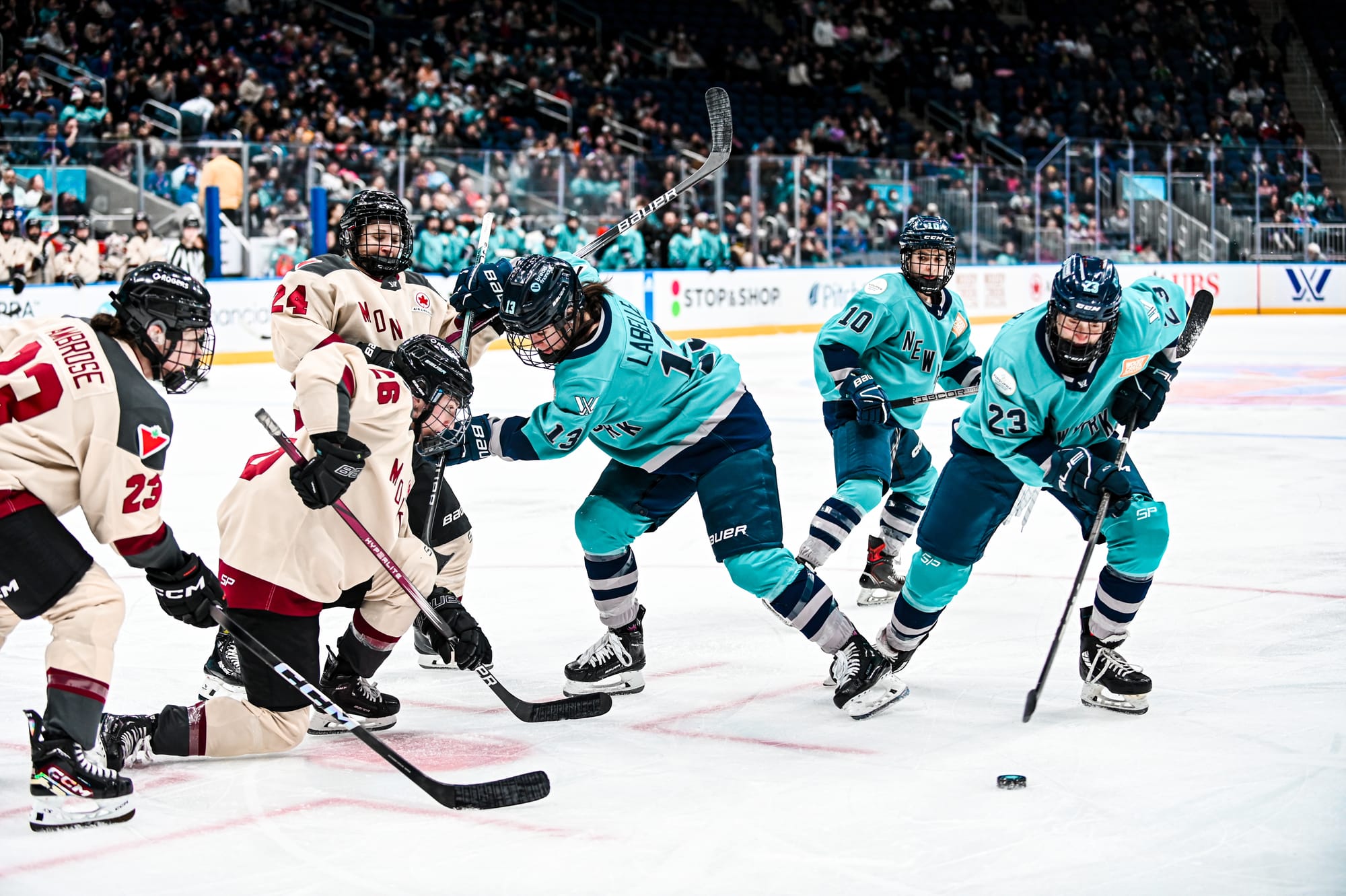 Madison Packer goes for the puck as other New York and Montréal players battle. The New York players are in teal home uniforms, while Montréal is wearing cream away uniforms.