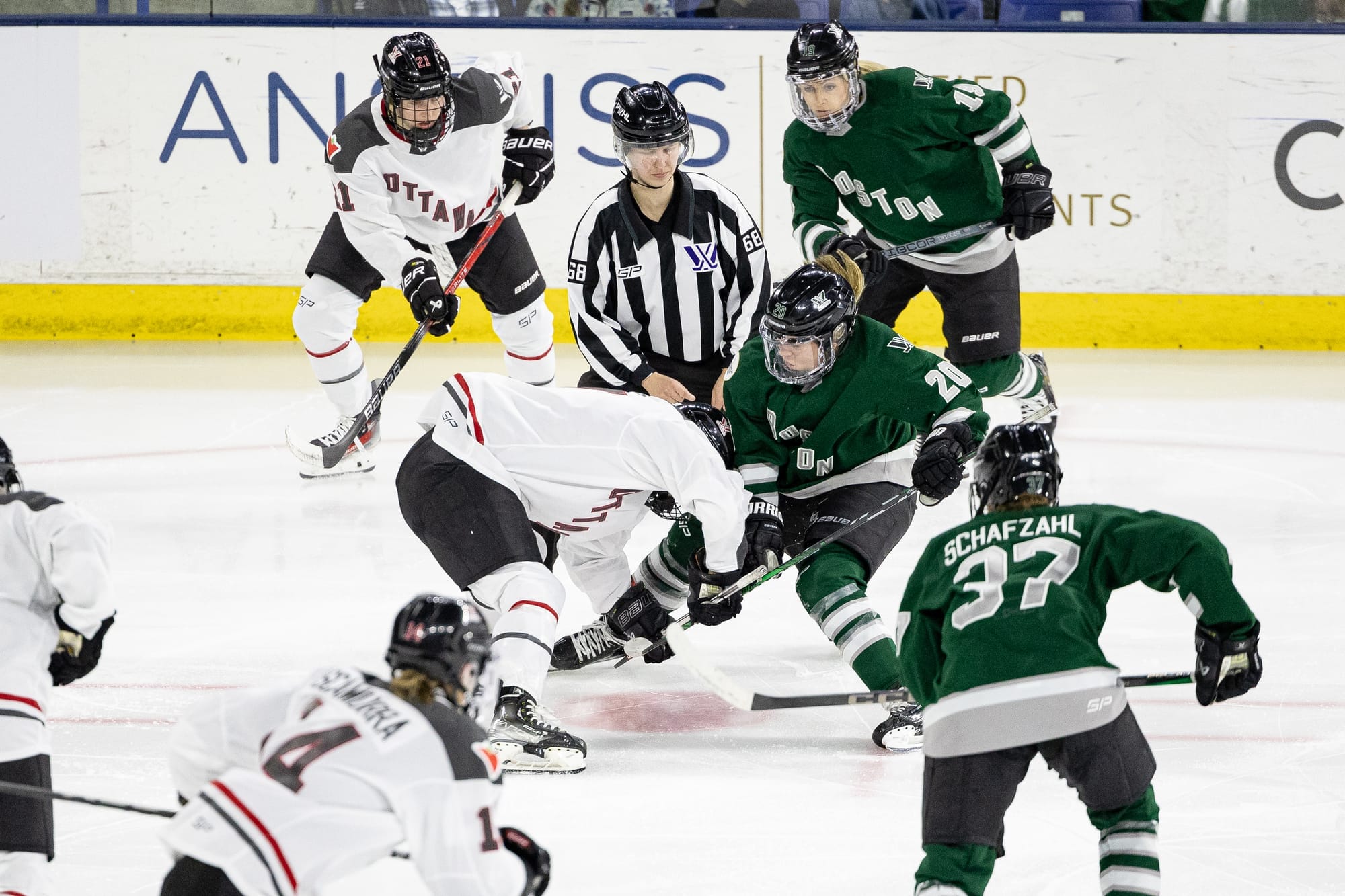 Hannah Brandt's line takes a face off against Ottawa. Boston is wearing green, while Ottawa is wearing white. A linesperson is standing in the middle of the two teams.