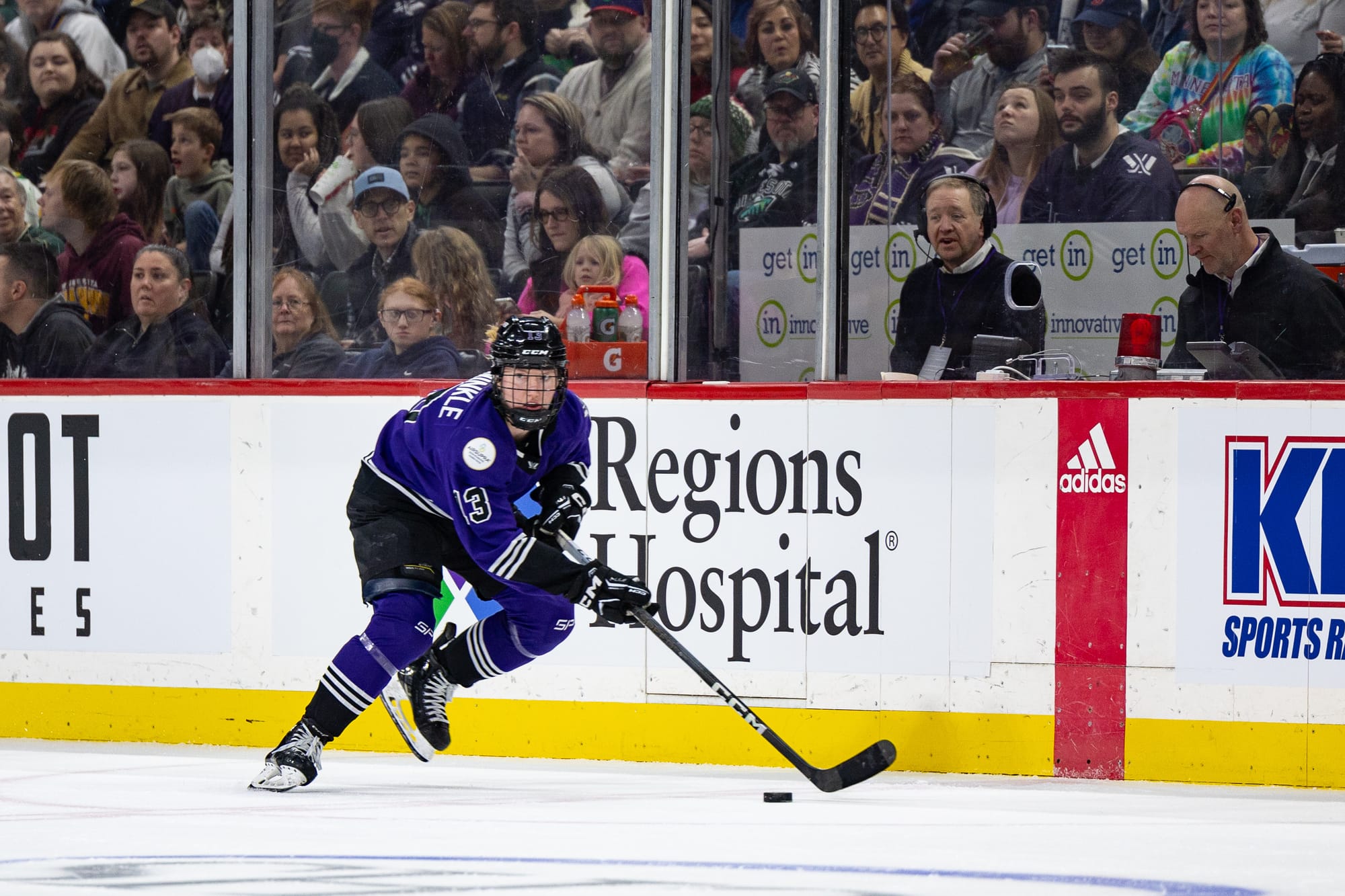 Grace Zumwinkle carries the puck during a game. She is bent over skating with the puck in front of her, and she is wearing her purple home uniform.