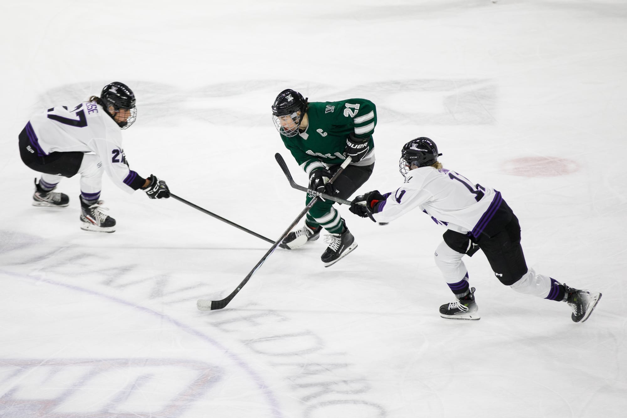 Hilary Knight skates the puck through Taylor Heise and Sophia Kunin. Knight is wearing a green home uniform, while Heise and Kunin and are in white away uniforms.