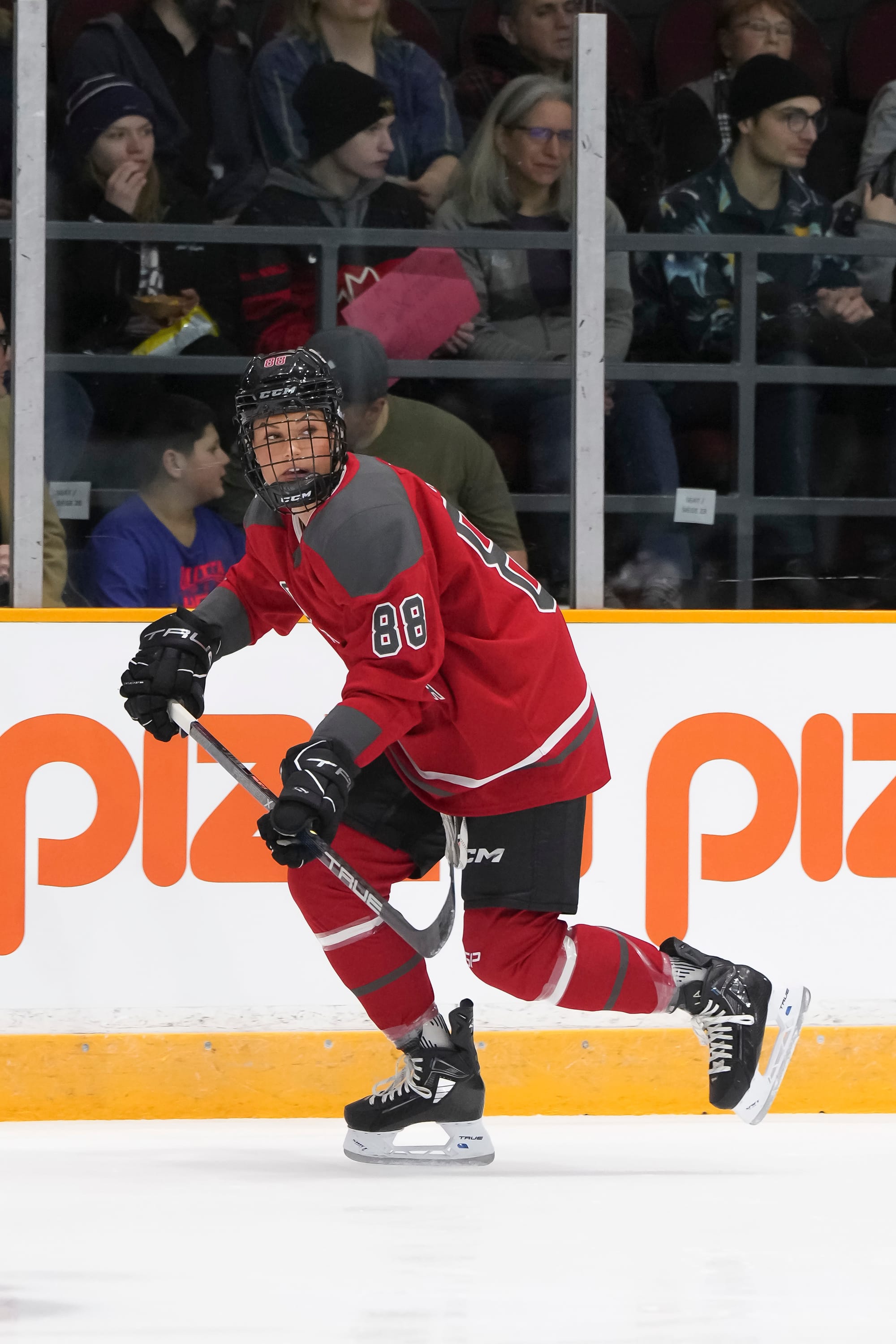 Lexie Adzija skates during a game. She is pushing off with her right foot while visually tracking the puck, and she is wearing her red home uniform.