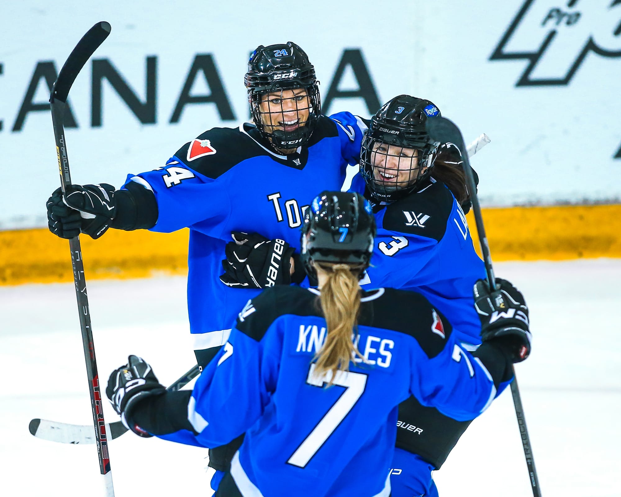 Natalie Spooner, Jocelyne Larocque, and Olivia Knowles celebrate a goal. They are in a group hug and all wearing blue home jerseys.