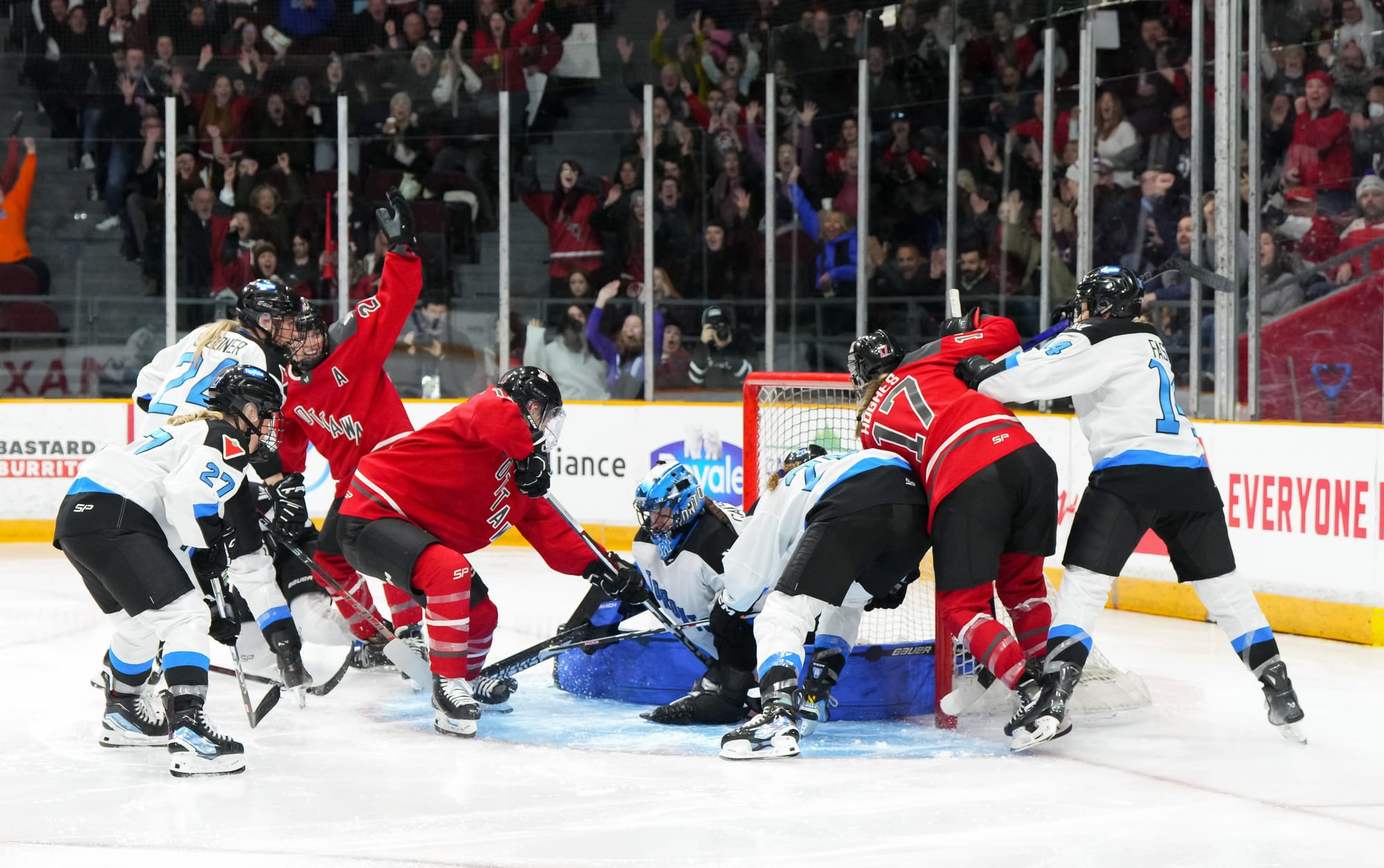 Ottawa players start to celebrate after scoring a scrappy goal. One has her arms raised while the other two are still battling by the goal crease. There are four Toronto players are them, plus goaltender Kristen Campbell. Ottawa is wearing red, Toronto in white.