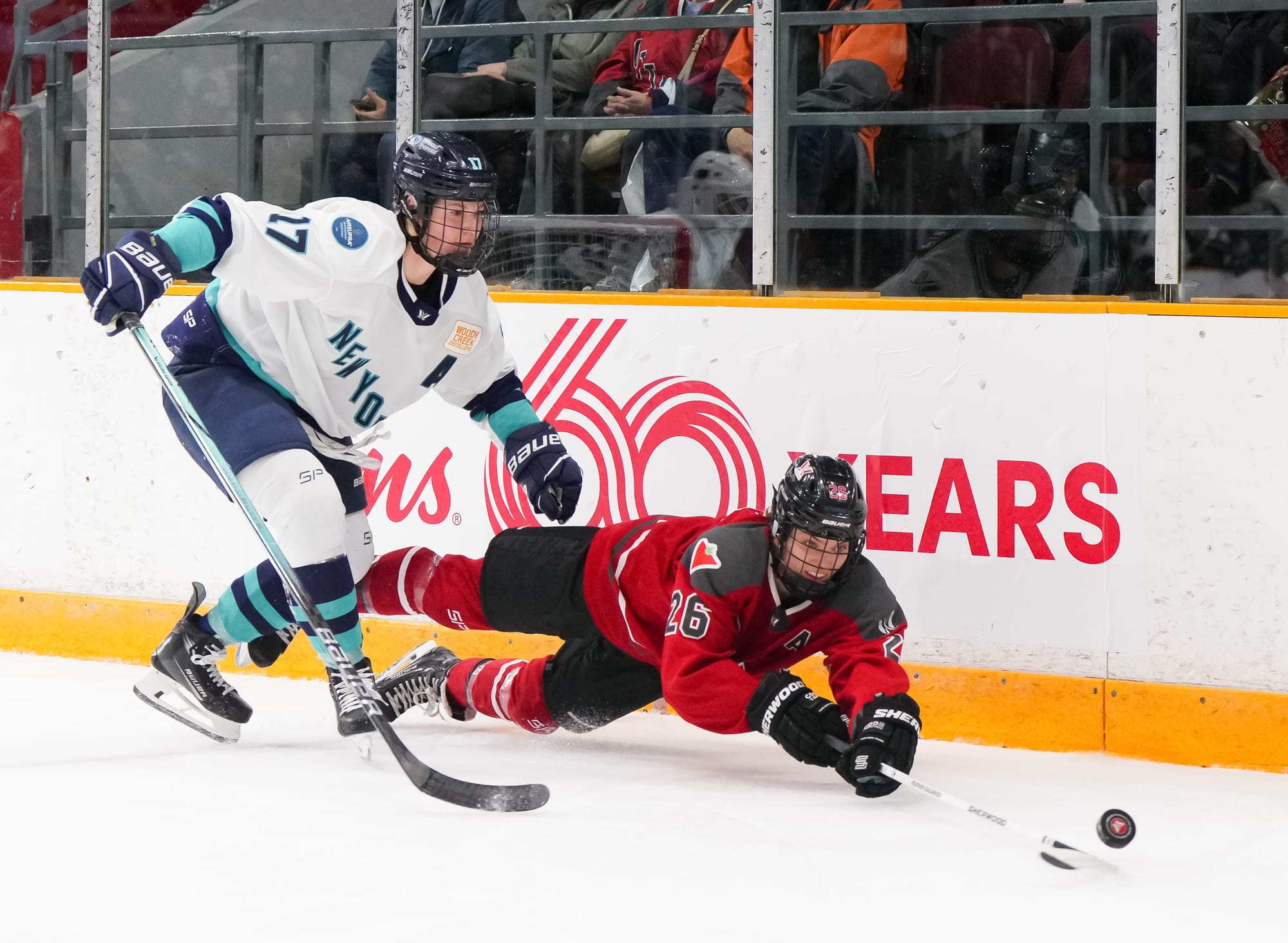 Emily Clark makes a play on the puck while falling, with Ella Shelton defending. Clark is nearly flat on the ice with her stick outstretched, while Shelton is still skating. Clark is in a red home uniform, while Shelton is in a white away.