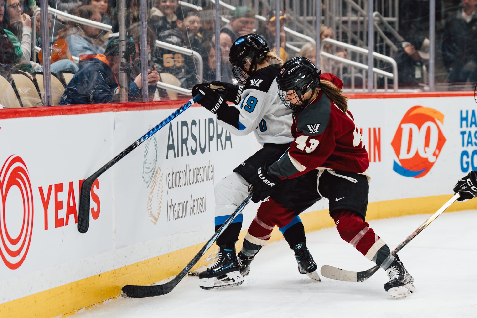 Kristen O'Neill battles behind the net with Rebecca Leslie. O'Neill is about to pin Leslie to the boards from behind in an attempt to retrieve the puck from between her feet. O'Neill is in maroon, while Leslie is in white.