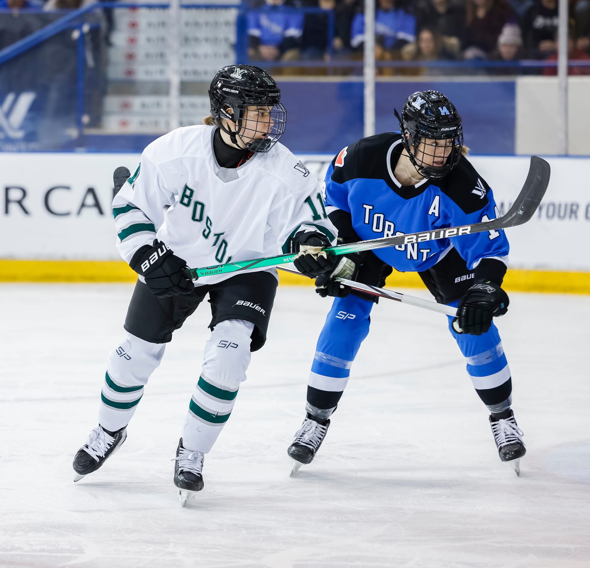 Alina Müller and Renata Fast hunt the puck. Müller is in white and is skating with her stick at waist-height. Fast is in blue and skating a bit hunched over with her stick also at wasit-heigh.