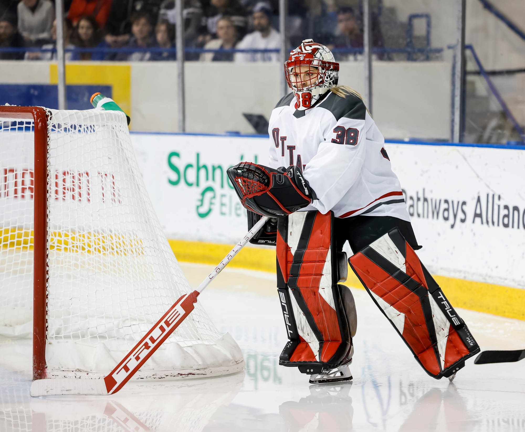 Emerance Maschmeyer skates back to her net after playing the puck during a game against Toronto. She is wearing her white away uniform, red/black/white leg pads, black/red gloves, and red/white mask.