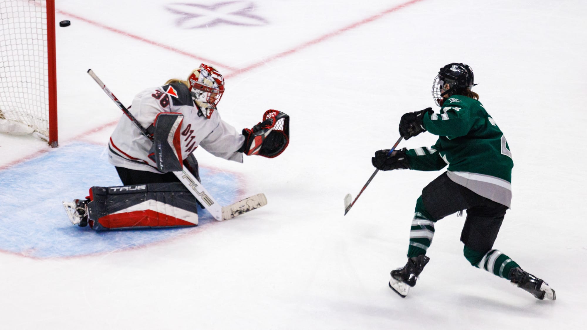 Hilary Knight scores on Emerance Maschmeyer. Knight is mid-follow through on the shot, with her weight primarily on her front (left) foot and stick in the air. Meanwhile, Maschmeyer is on her knee with her glove outstretched low as the puck sails high over it. Knight is in green, while Maschmeyer is in white. 