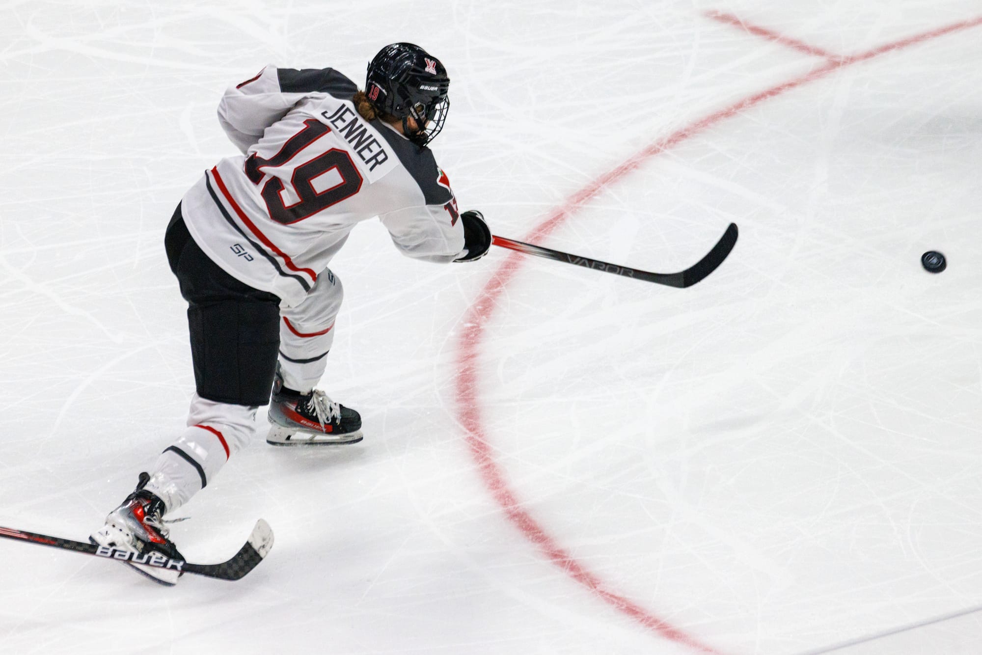 Brianne Jenner takes a shot against Boston. The photo is taken from behind. She has her weight on her left (front) foot as she follows through on the shot. The puck is just above to leave the frame. Jenner is wearing white.