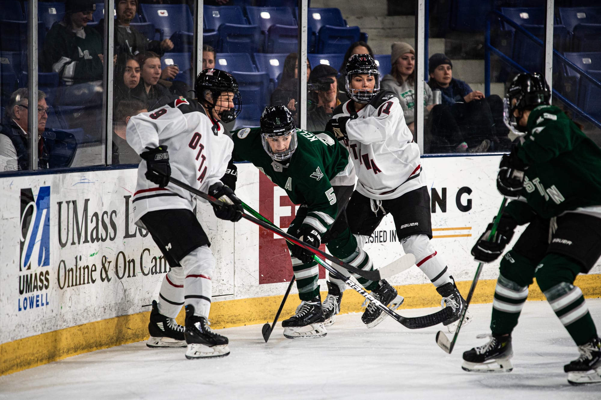 Megan Keller fends off two Ottawa players in the corner. Keller is in the middle, hunche over, while the Ottawa players are upright. Keller is wearing her green home uniform, while Ottawa is in white.