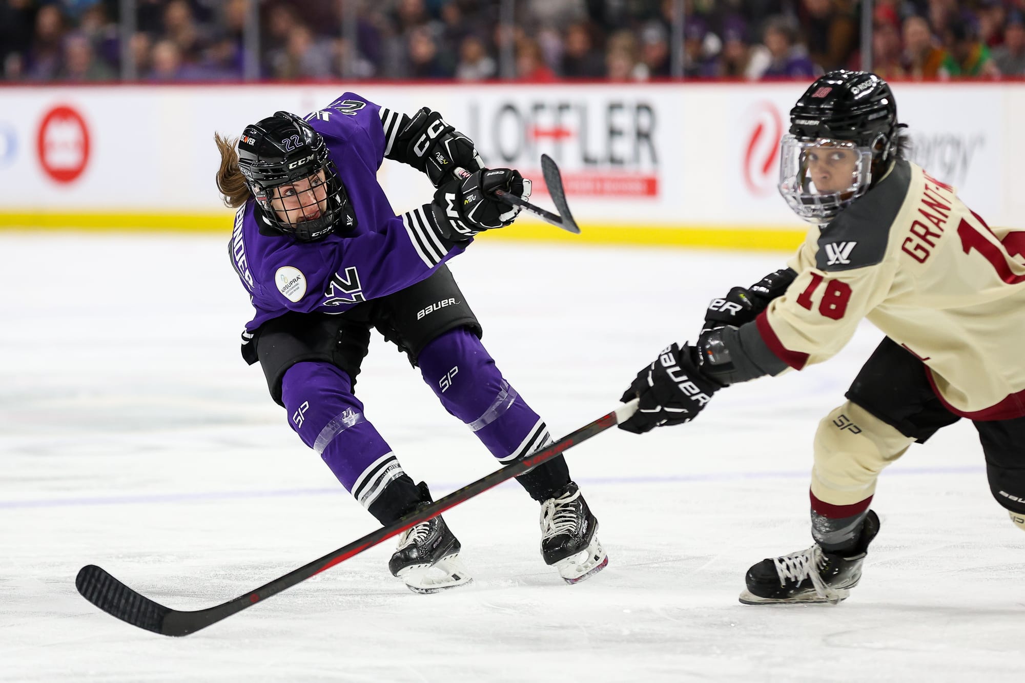 Natalie Buchbinder takes a shot against Montréal as Mikyla Grant-Mentis defends. Buchbinder is hunched over with her stick raised in a follow-through. Grant-Mentis is hunched over with her stick on the ice outstretched to try to defend. Buchbinder is in purple, Grant-Mentis is cream.
