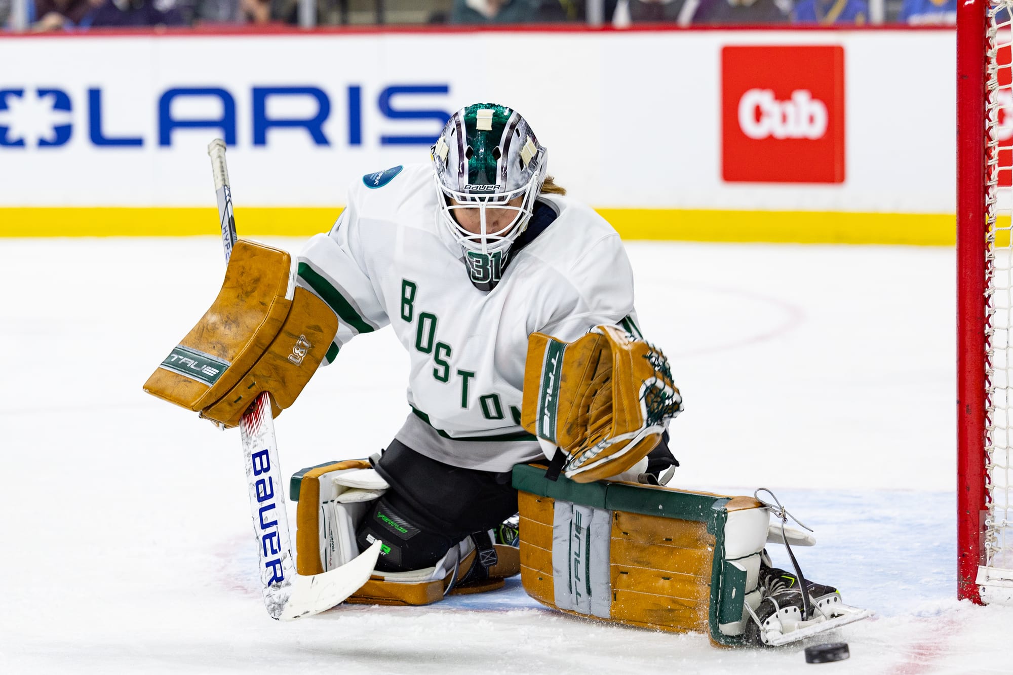 Aerin Frankel makes a save against Minnesota. She is on her knee and kicking the puck out to her left. She is in her brown pads, green and white mask, and white away uniform.