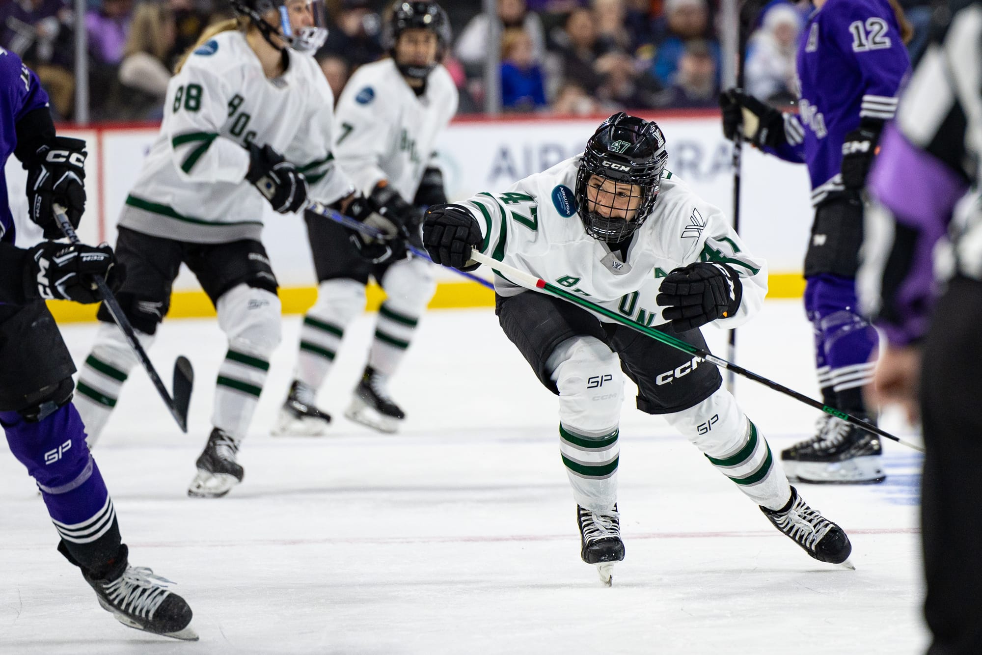 Jamie Lee Rattray hunts the puck against Minnesota. She is hunched over skating, with one hand outstreched and holding her stick. She is in a white away uniform.