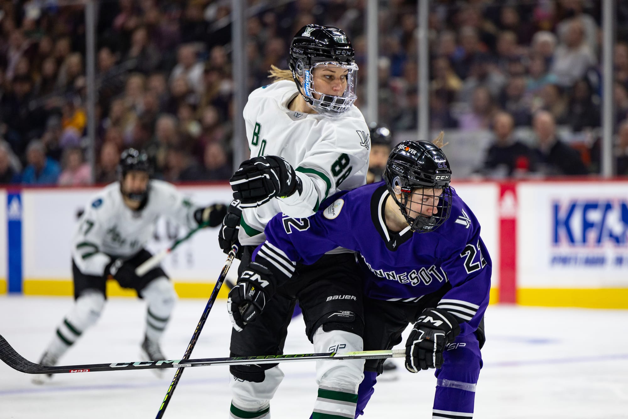 Susanna Tapani and Natalie Buchbinder battle in pursuit of the puck. Buchbinder is hunched over pushing in to Tapani, while Tapani is upright and trying to get around Buchbinder. Tapani is wearing a white away uniform, while Buchbinder is in a purple home one.