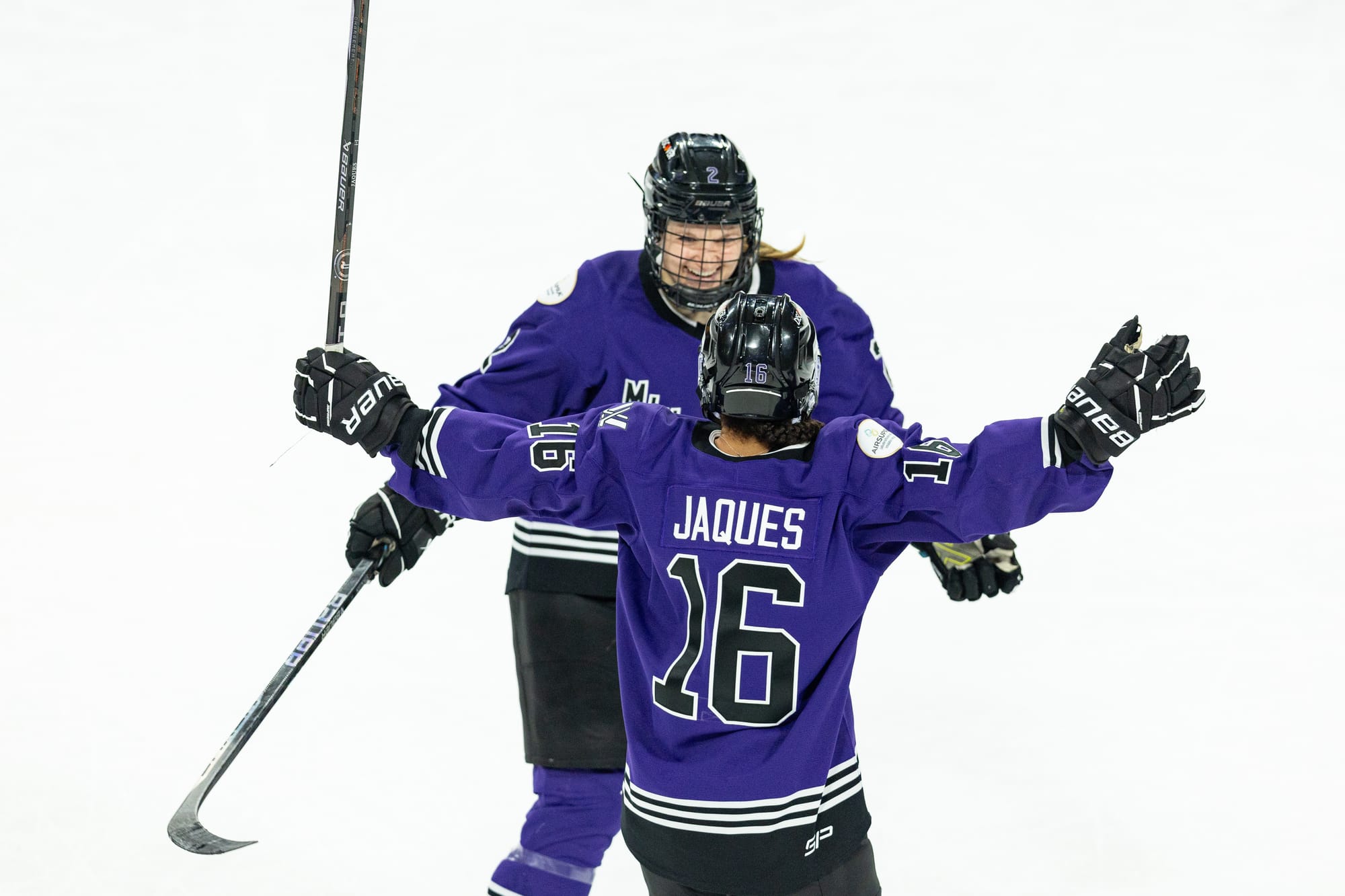 Sophie Jaques celebrates one of her goals against Toronto with Lee Stecklein. Jaques has her arms up and open, waiting for Stecklein to arrive for a hug. Both are in their purple home uniforms.