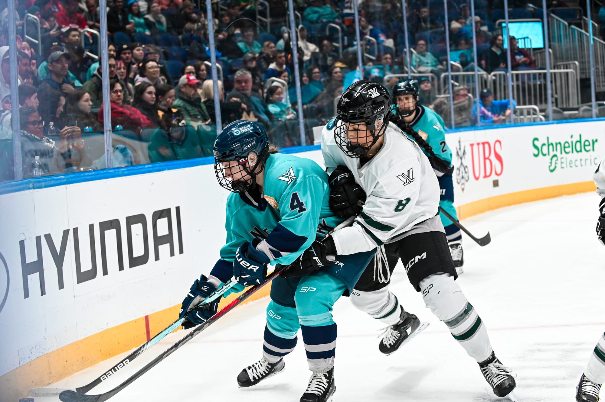 Taylor Baker tries to protect the puck from Lexie Adzija. Baker is hunched over further into the corner, as Adzija reaches around her. Baker is in a teal home uniform, while Adzija is in a white away uniform.