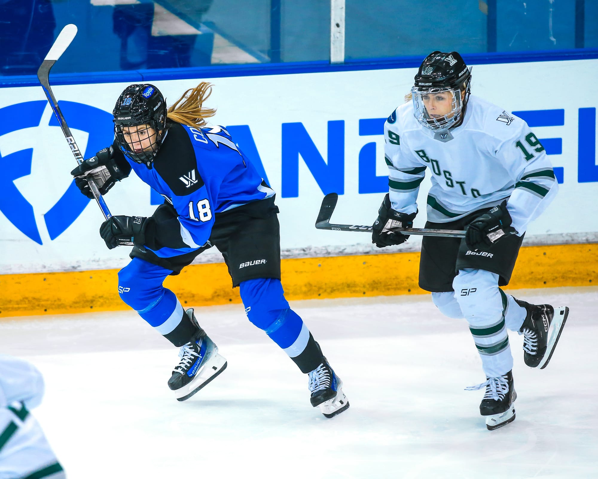 Gigi Marvin and Jesse Compher hunt the puck. Compher is slightly ahead of Marvin. Both are slightly hunched over while skating. Marvin is wearing white, while Compher is wearing blue.