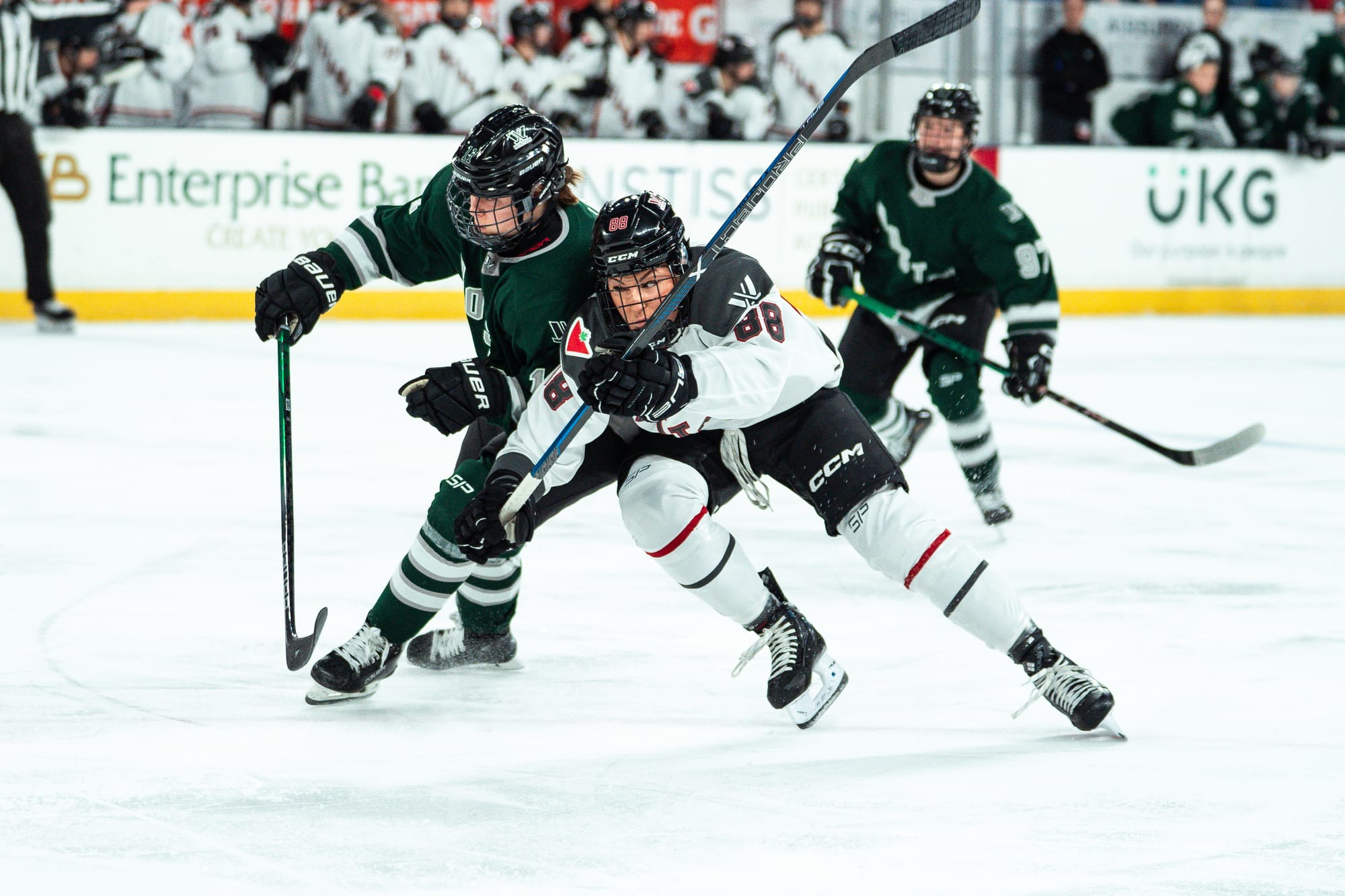 Alina Müller and Lexie Adzija battle for positioning as they hunt the puck. They are leaning into each other and both hunched over, with Müller on the left. Müller is in her green home uniform, while Adzija is in white.