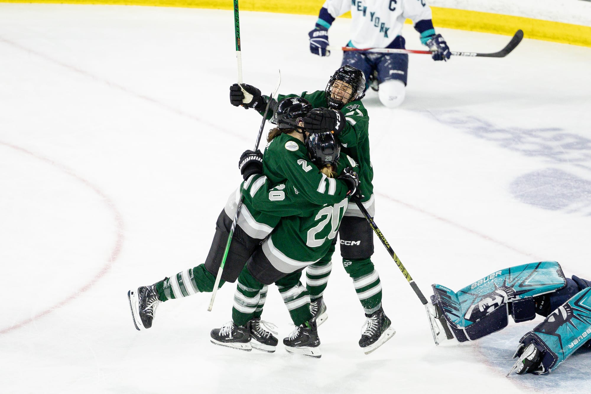 Emily Brown and Jamie Lee Rattray hug-tackle Hannah Brandt following her OT winner. Each player is wearing their green home jersey, and Brandt is starting to fall over. In the bottom right corner are Abbey Levy's legs as she lays on the ice after allowing the goal.