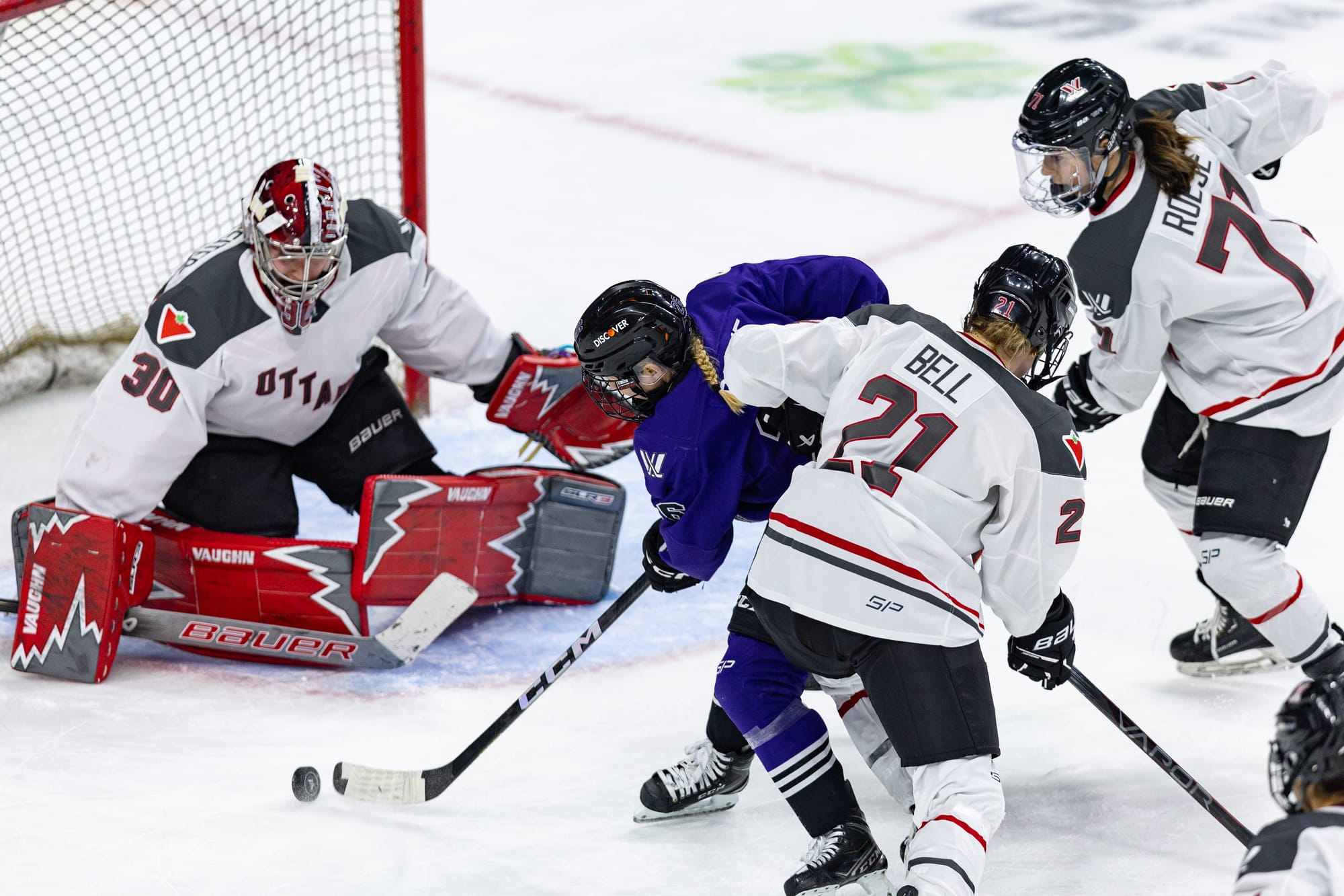 Sandra Abstreiter makes a save on Kendal Coyne Schofield as Ottawa players try to defend. Abstreiter is on her knees while Schofield is hunched over ready to shoot. There are two Ottawa players behind her. Ottawa is in white, while Schofield is in purple. Abstreiter is also wearing red and black pads and a red and white mask.