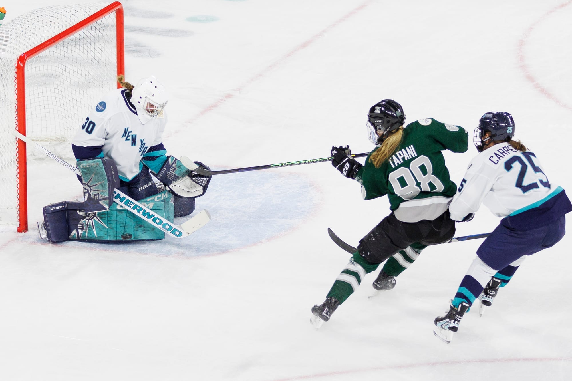 Susanna Tapani takes a shot in close on Corinne Schroeder. Alex Carpenter is right behind Tapani trying to defend. Tapani is in green, while Schroeder and Carpenter are in white.