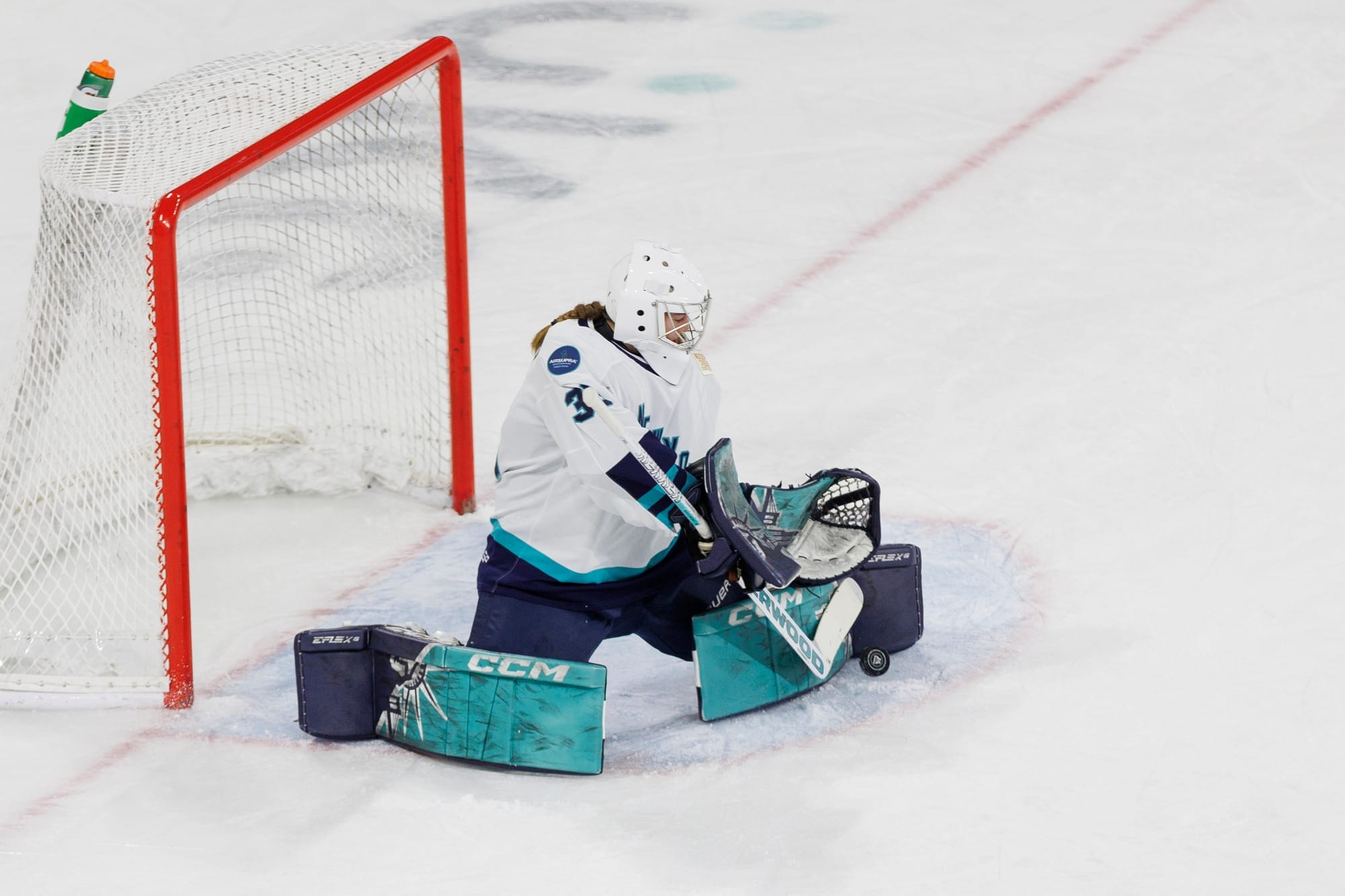 Corinne Schroeder makes a save against Boston. She is on her knees and kicking the puck out to her left. She is wearing a white mask and jersey to go with her teal/navy New York pads.