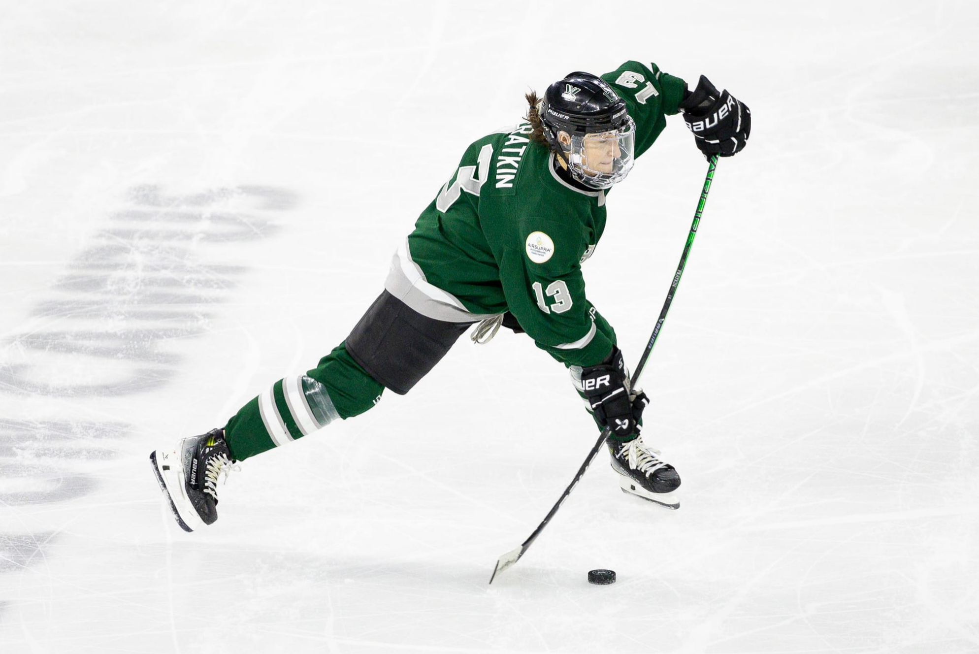 Kaleigh Fratkin moments before taking a slap shot against New York. Her weight is on her left leg, which is out front, while her stick is flexed as it's about to hit the ice before following through on the puck. She is wearing a green home uniform.