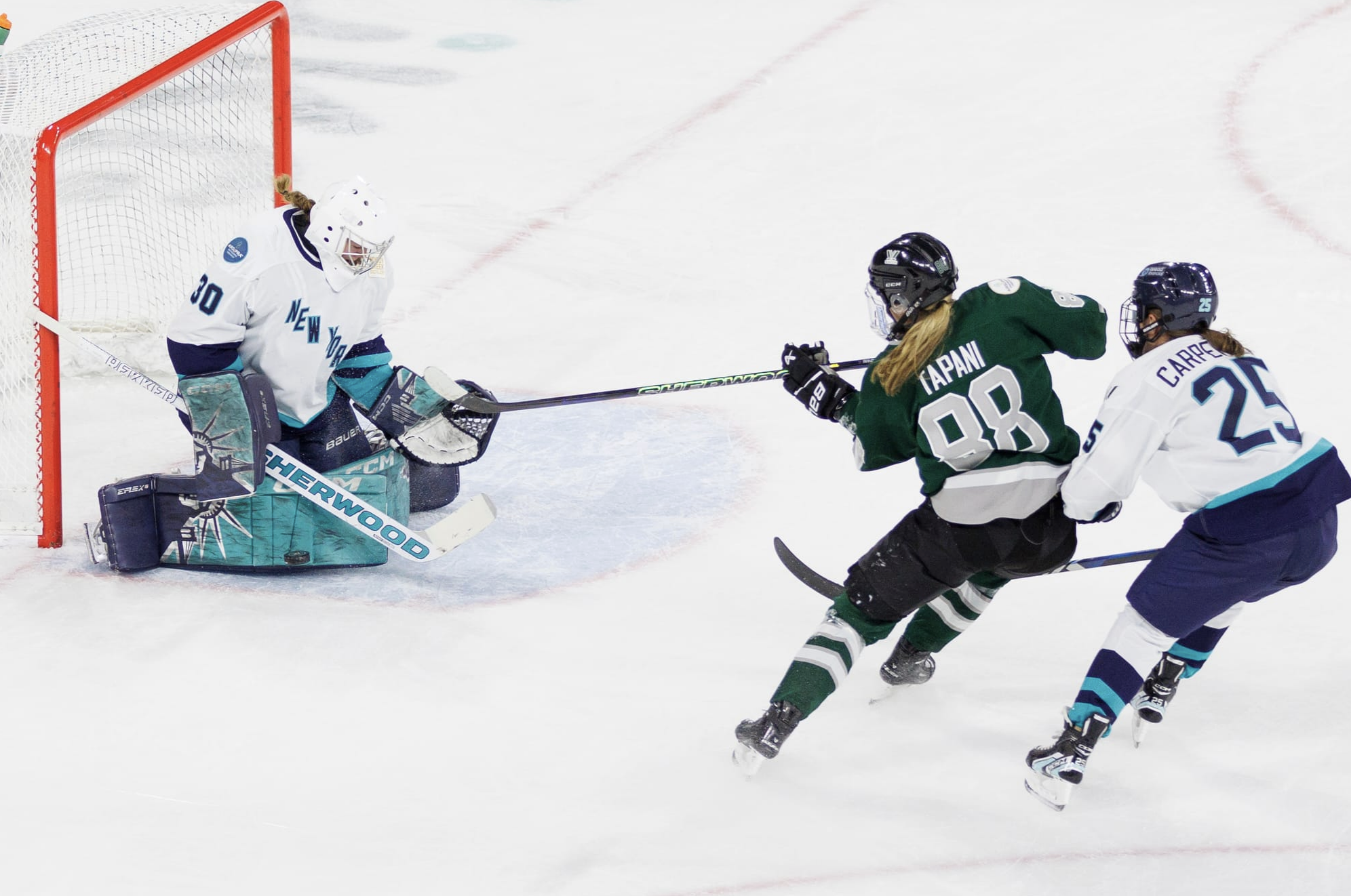 Schroeder makes a save on Tapani. Tapani's stick is raised for the shot follow through while Alex Carpenter defends. Meanwhile, Schroeder is on her knees squeeze her pads together for the save. Tapani is in green, while the New York players are in white. 