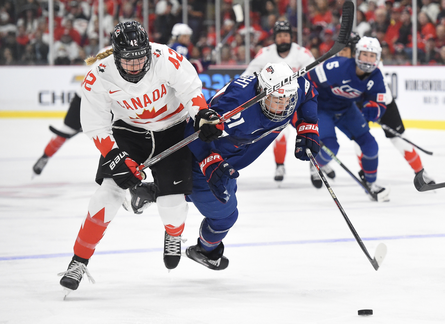 Hilary Knight falls as Claire Thompson battles her for the puck. Knight is mid-air, and she was carrying the puck but appears about to lose it as she falls forward. Meanwhile, Thompson is still upright and her stick is in the air as she moves in to take the puck. Thompson is in white, while Knight is in navy.