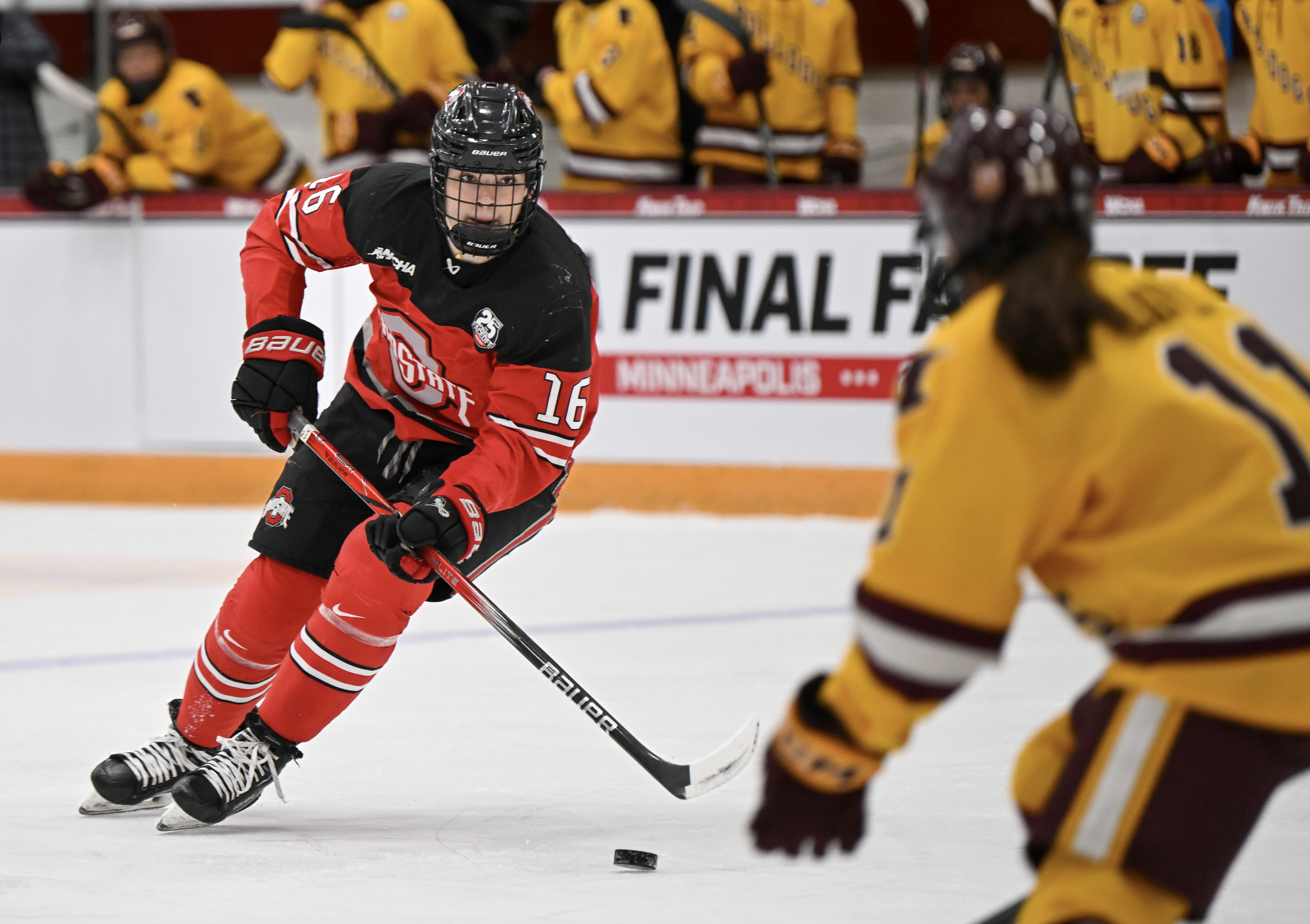 Joy Dunne skates with the puck. She is hunched over amd looking up at a defender, who is blurry in the shot. Dunne is wearing a red and black OSU uniform.