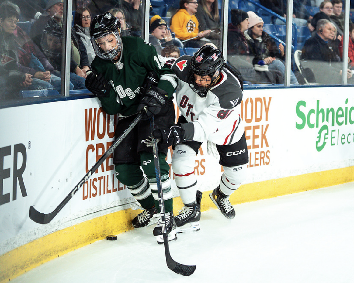 Adzija body checks Marvin behind the net as they battle for the puck. Adzija is wearing a white Ottawa uniform, while Marvin is in a green Boston uniform.