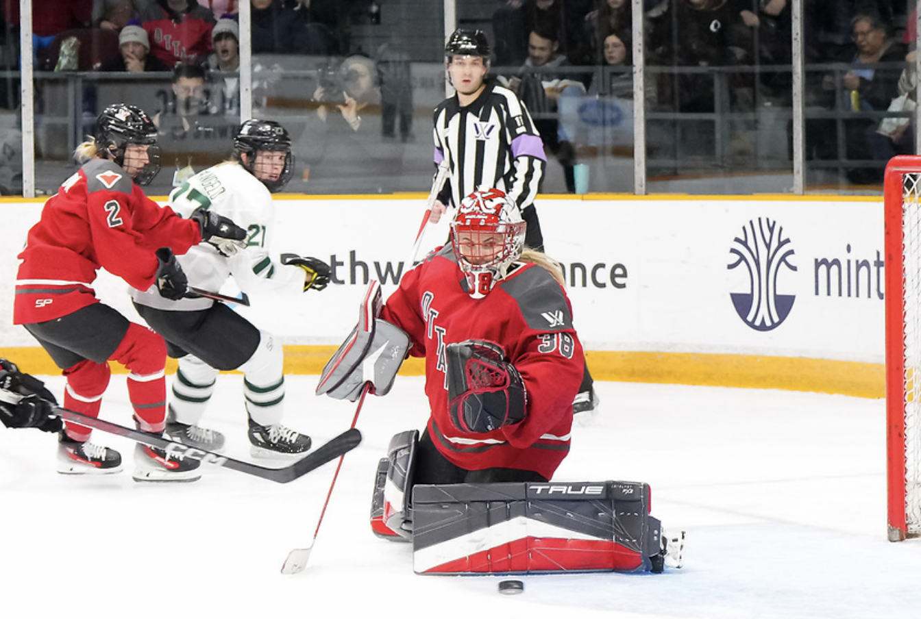 Maschmeyer makes a save against Boston from the butterfly position. She is kicking the puck to her left, as an Ottawa defender pushes a Boston player out of the play. The Ottawa players are wearing red home uniforms, while the Boston player is wearing a white away uniform. 