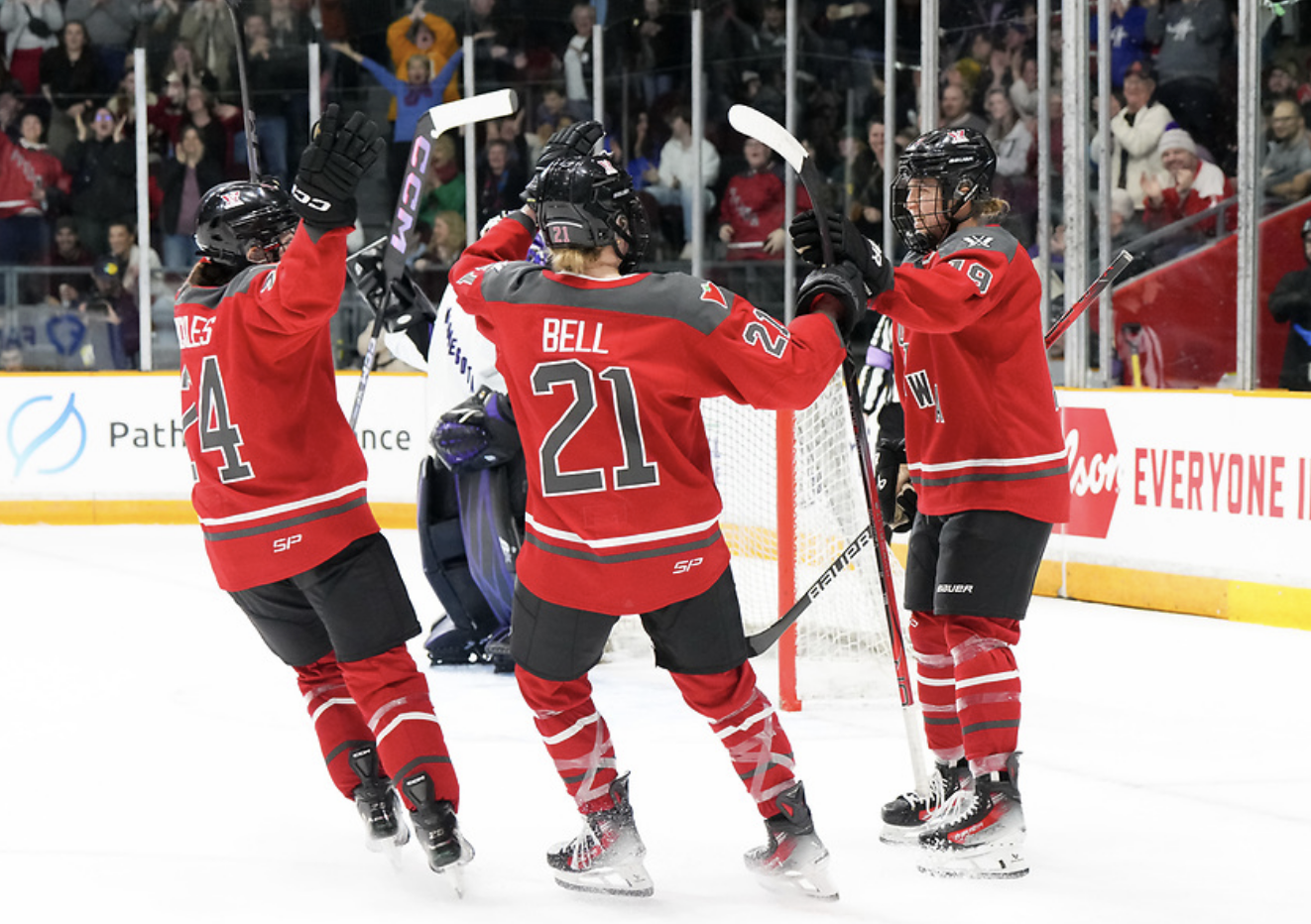 Emma Buckles and Ashton Bell skate to hug Brianne Jenner after one of her goals against Minnesota. Jenner is pointing at them, while Bell and Buckles have their arms raised. They're all in red home uniforms.