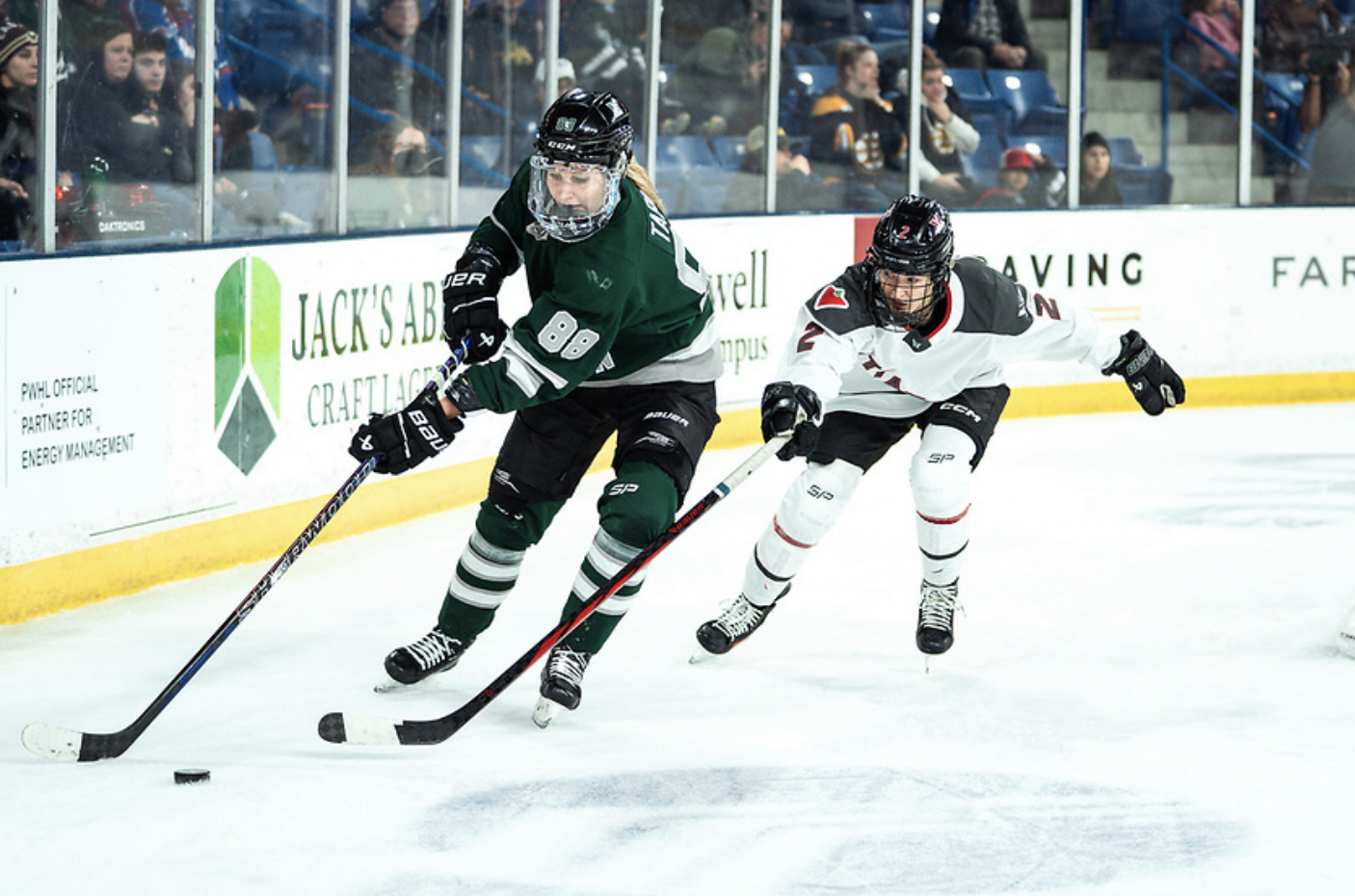 Tapani leans toward the trailing and reaching Tejralová to shield the puck from her. Tapani is wearing a green home uniform, while Tejralová is in a white away uniform. 