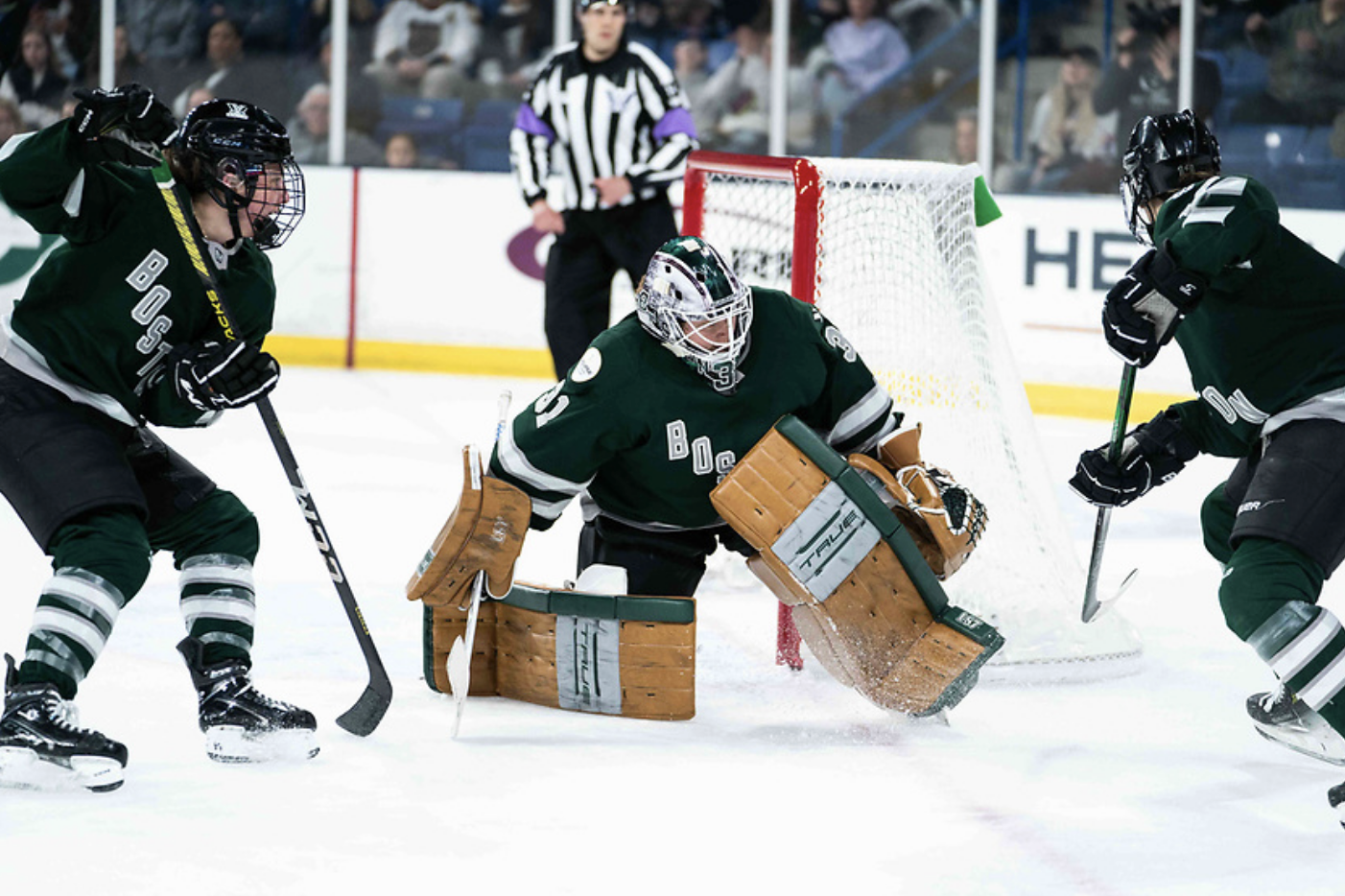 Frankel, getting up from her knees, resets after making a save as two teammates on either side of her search for the puck. All are in green home uniforms.