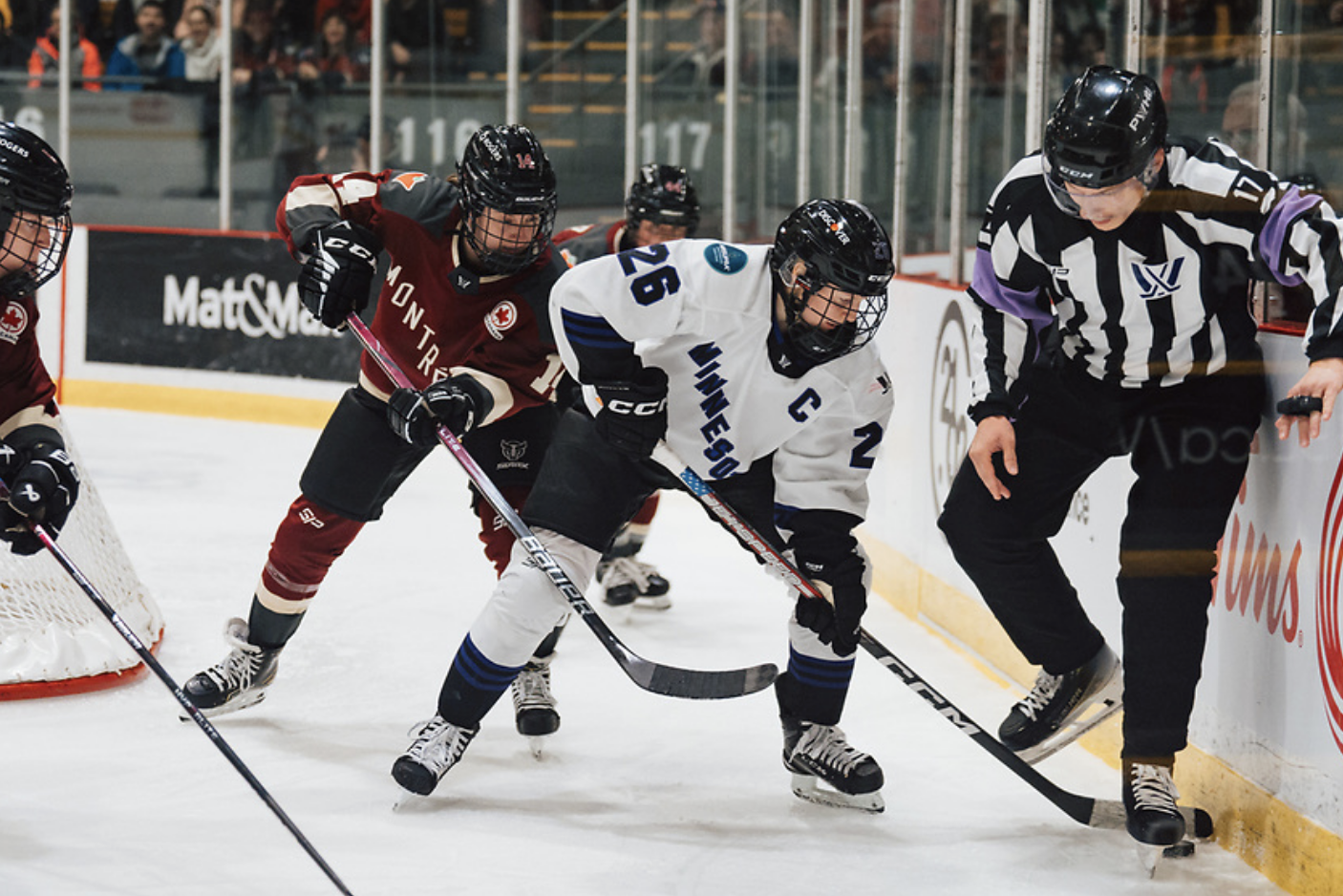 Coyne Schofield goes to play the puck behind the net while under pressure from Montréal's Jillian Dempsey. The puck is currently stuck between a referee's feet. Coyne Schofield is in white, while Dempsey is in maroon.