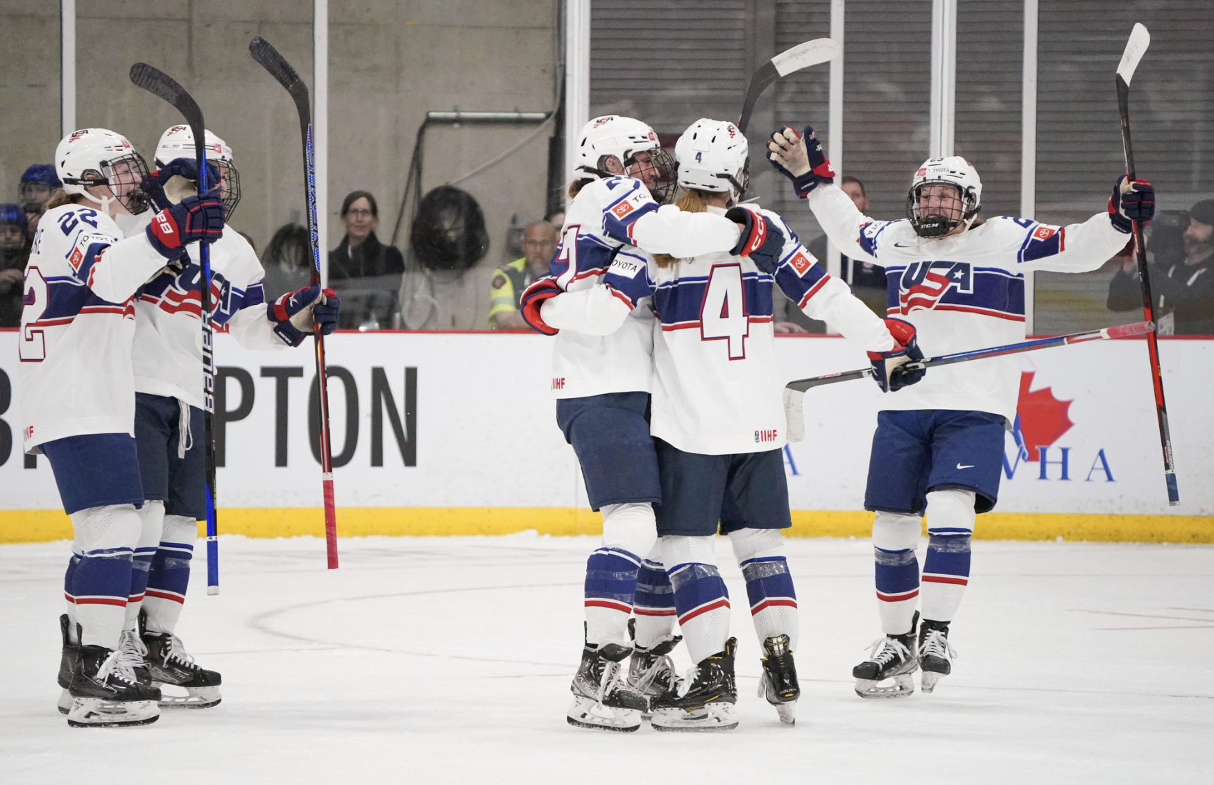 Team USA players celebrate a goal against Switzerland with a group hug. Two are already in the hug, while two others skate from the left to join and one from the right. They're all wearing white USA uniforms.