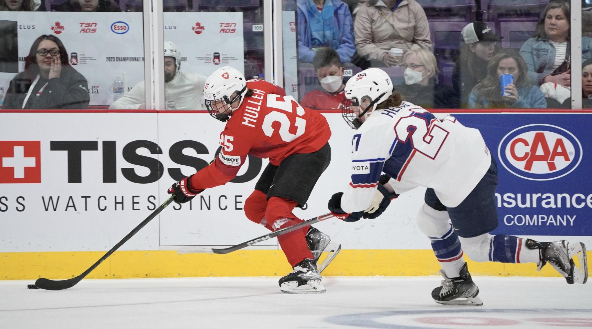 Alina Müller skates with the puck as Taylor Heise tries to catch her. Heise's stick is outstretched as she goes to defend. Müller is in a red uniform, while Heise is in white.