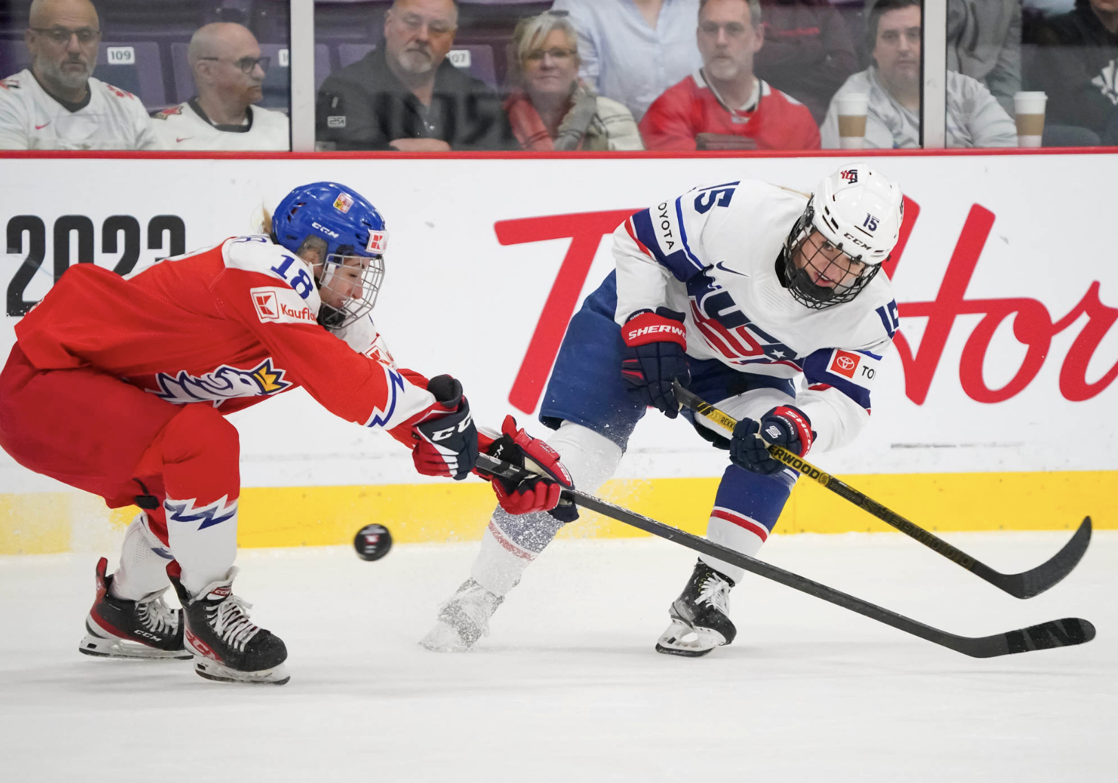 Savannah Harmon (right) makes a pass under Michaela Pejzlová's stick. Both players are hunched over with outreached sticks. Harmon is wearing white, Pejzlová is in red.