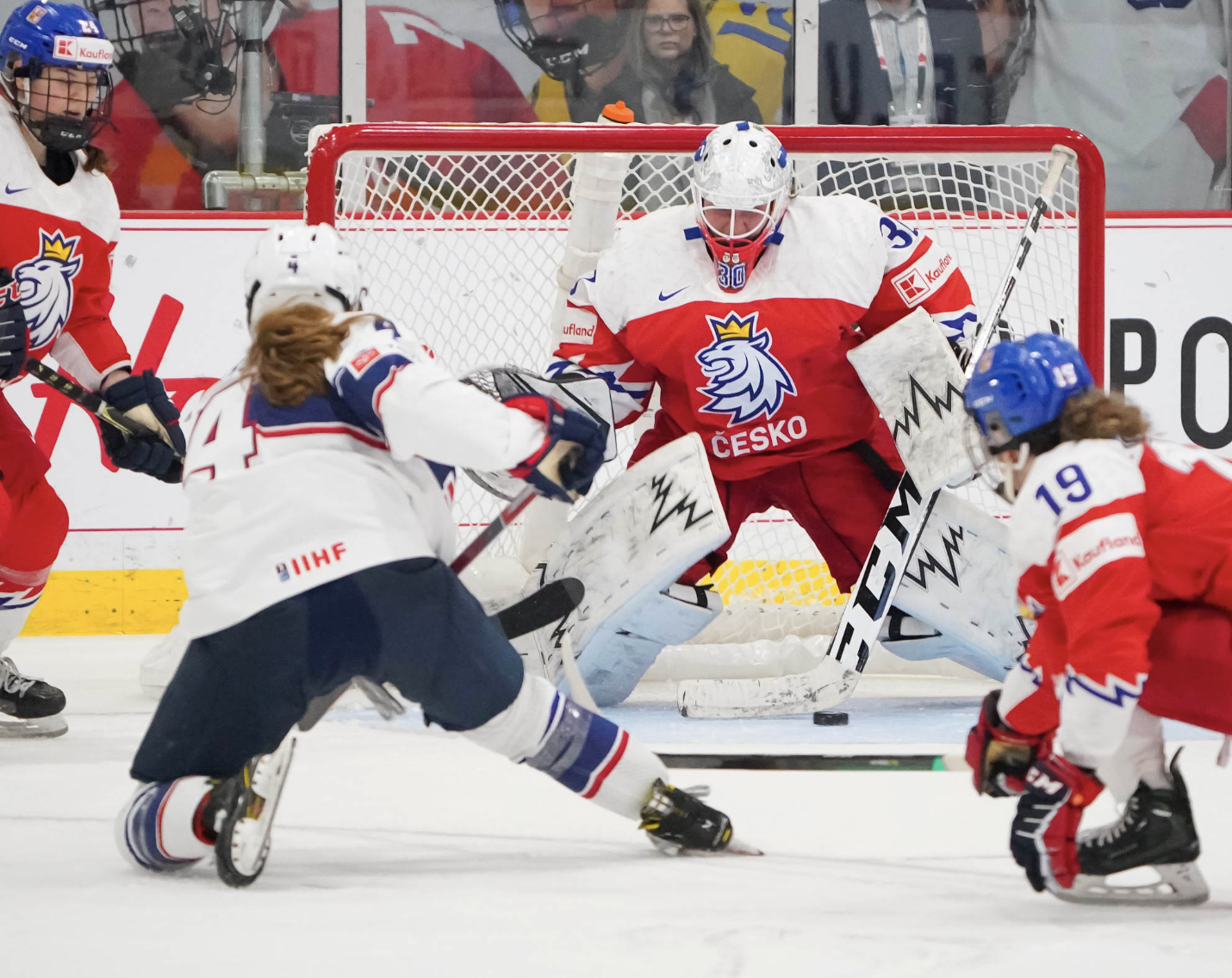 Harvey takes a shot on Zechovská. Harvey is on her knee following the shot, while Zechovská is dropping down to hers while making the save. Two Czech defenders are visible on the edges of the photo. Harvey is wearing white, while all the Czechs are in red.