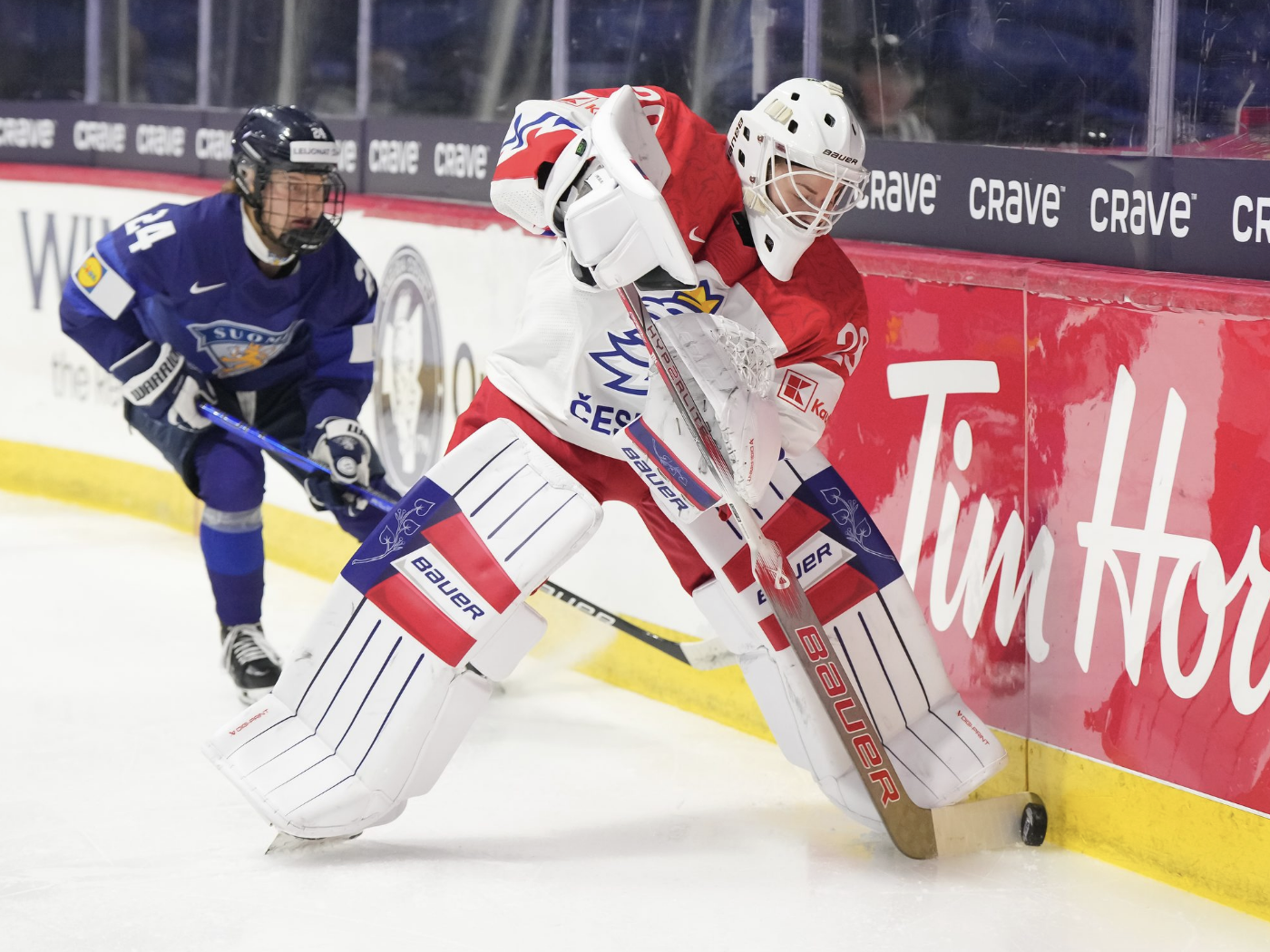 Klára Peslarová makes a play on the puck behind her net. There is a Finnish defender in pursuit. Peslarová is wearing a white uniform with red shoulders, while the Finnish defender is wearing a navy blue uniform.