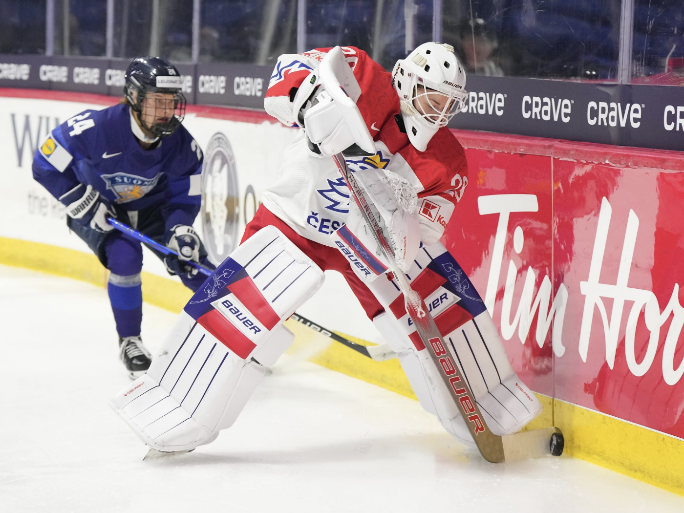 Klára Beslarová rozehrává puk za svou sítí.  Následuje ochranka z Finska.  Beslarová nosí bílou uniformu s červenými rameny, zatímco finský obránce nosí námořnickou modrou uniformu.