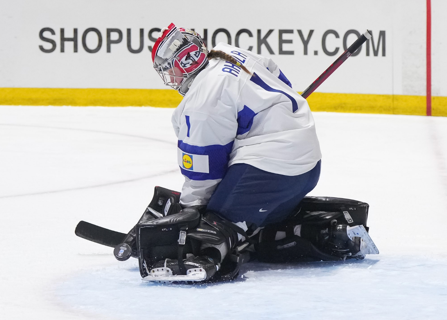 Ahola makes a save against Canada. She is on her knees and kicking the puck out to her left. She is wearing her St. Cloud State mask and white Finland uniform to go along with black pads.