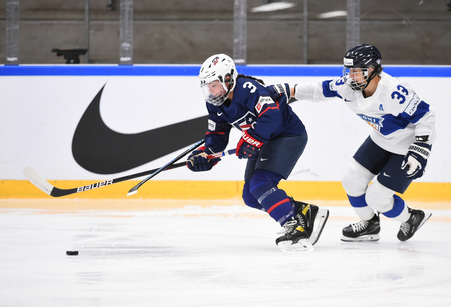 Barnes skates with the puck while Karvinen defends. Barnes is in the lead, hunched over with her stick outstretched and the puck in front of her. Karvinen is hunched over skating and reaching around Barnes with her stick. Barnes is in navy blue, while Karvinen is in white.