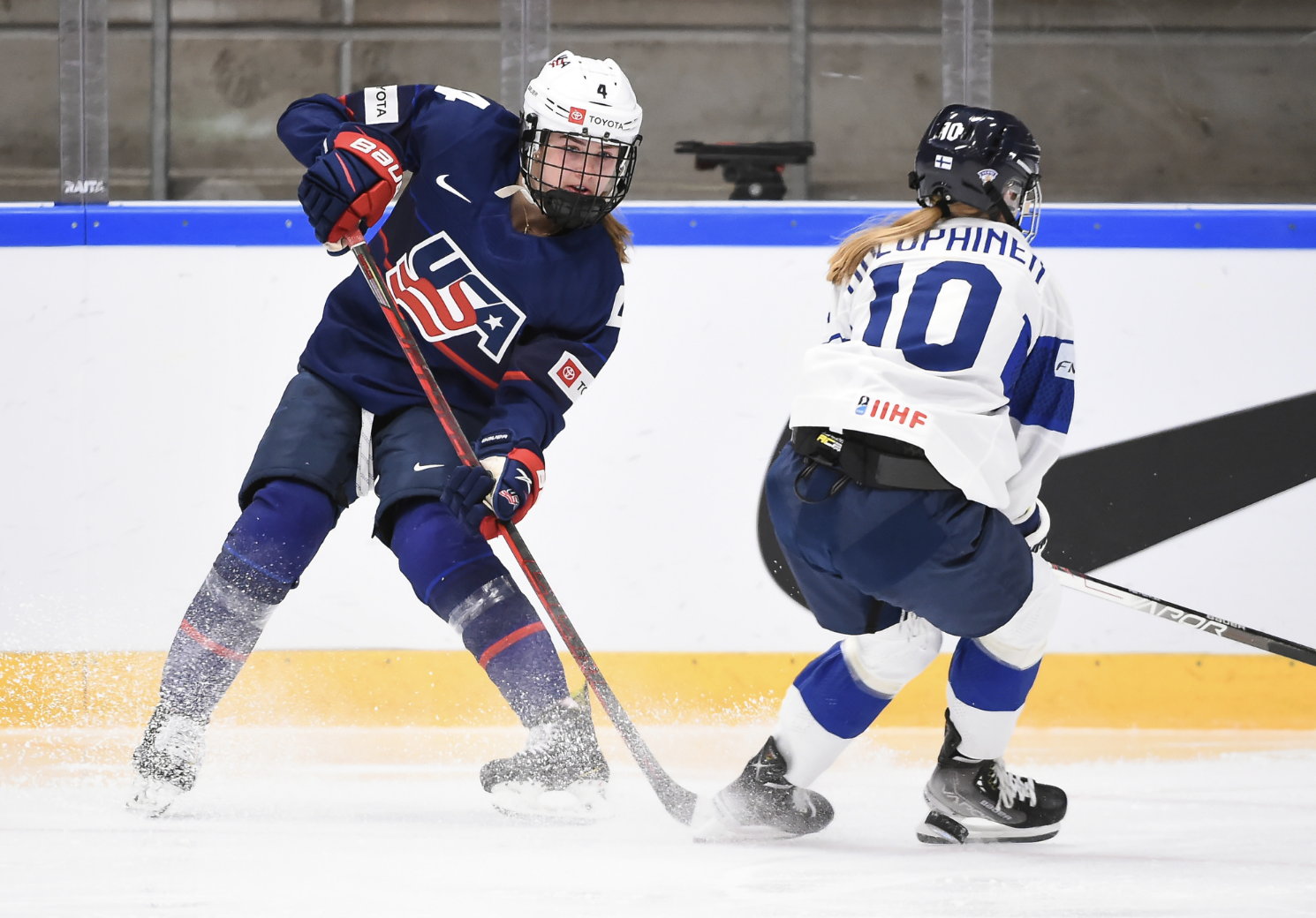 Harvey (left) dishes the puck as Holopainen (right) tried to block it. Harvey is hunched over making the pass, while Holopainen is nearly in a sitting position as she comes to a stop. Harvey is in navy blue, while Holopainen is in white