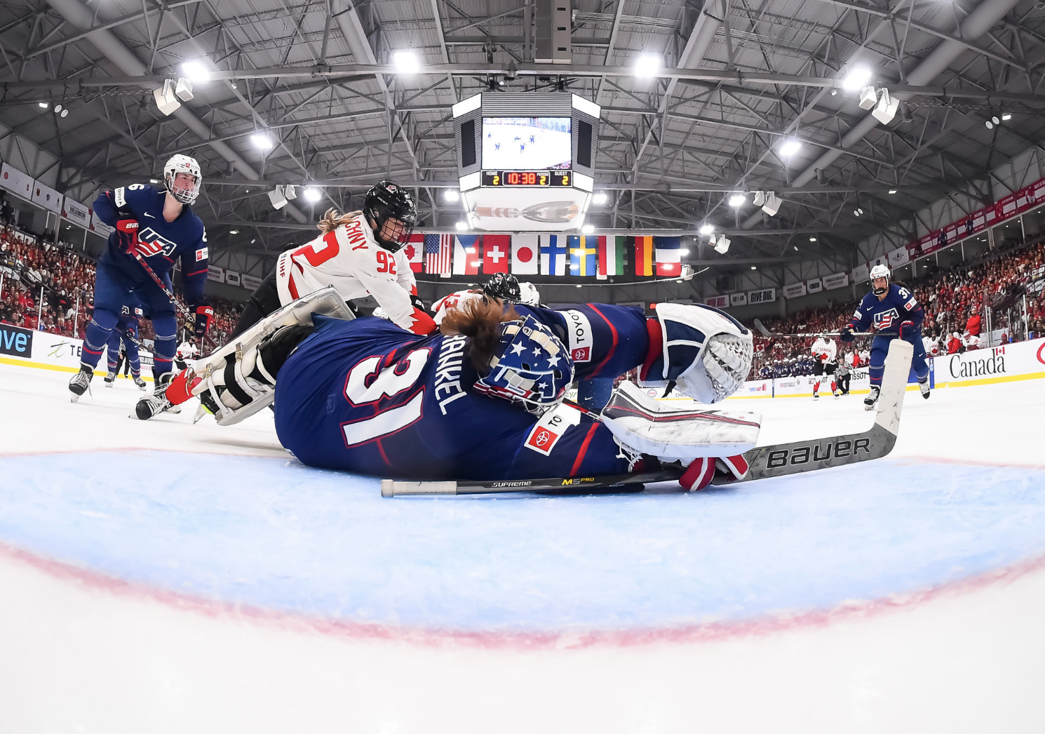 Frankel lays out to make a save against Canada. The shot is taken from inside the net, so it's of Frankel's back. She is wearing navy and laying with arms and legs outstretched, and the puck is just visible under her stick. Multiple skates from each side are in the background.