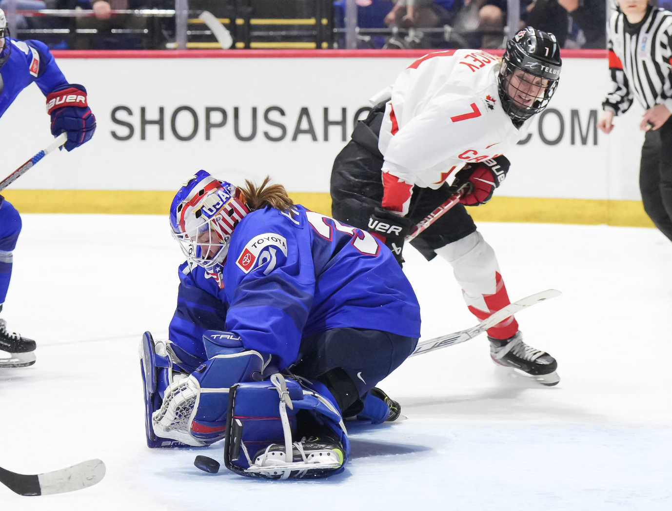 Frankel makes a save from the butterfly position. Her hands are in front of her, and the puck is next to her left leg. Her stick is caught up with Stacey, who is going past Frankel to her right after taking the shot. Frankel is in blue, while Stacey is in white.