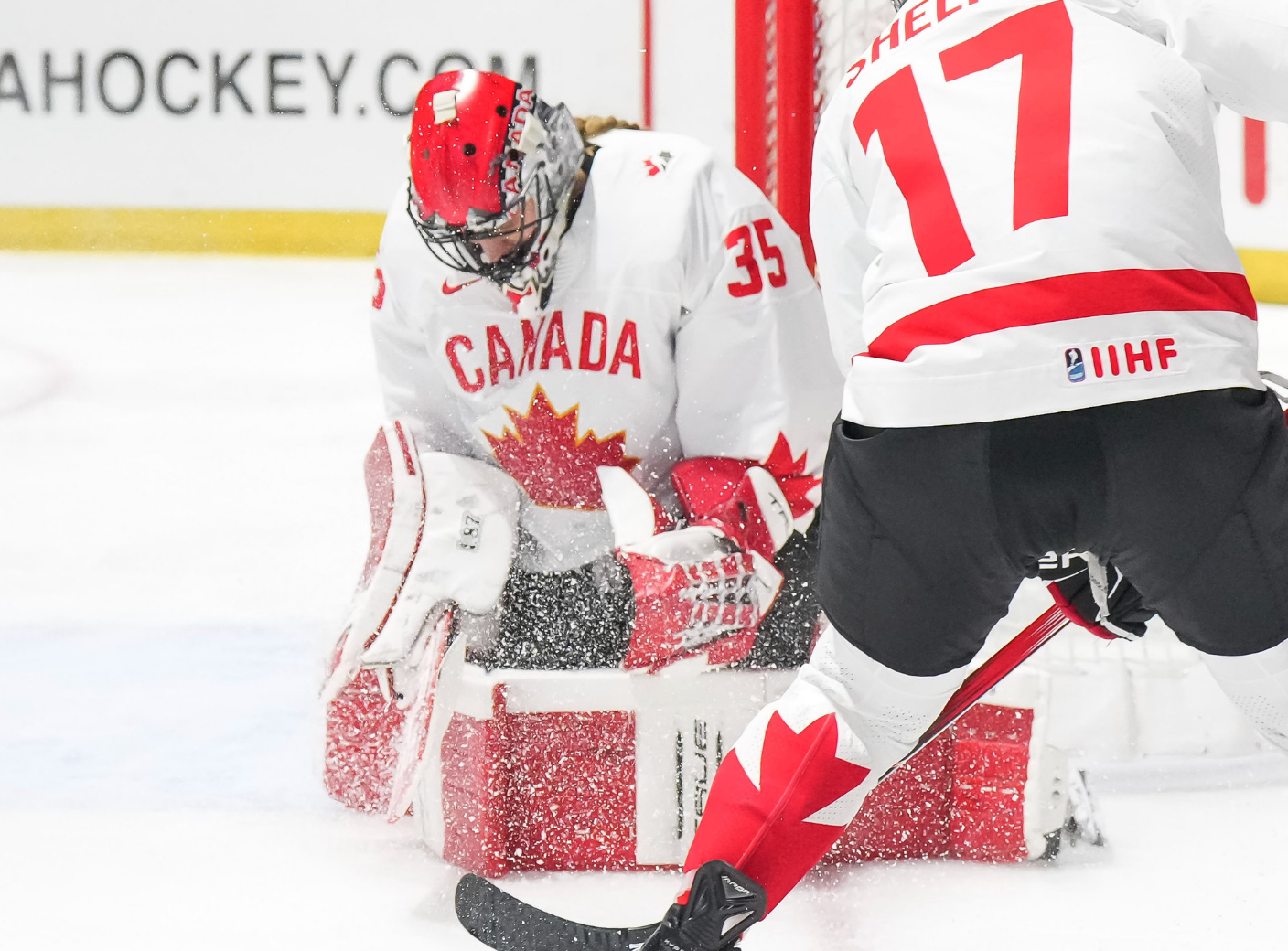 Desbiens squeeze her pads together to make a save on her knee. Her arms are pinned to her sides. Shelton is off to the side facing her. Both are in white and red Canada uniforms.